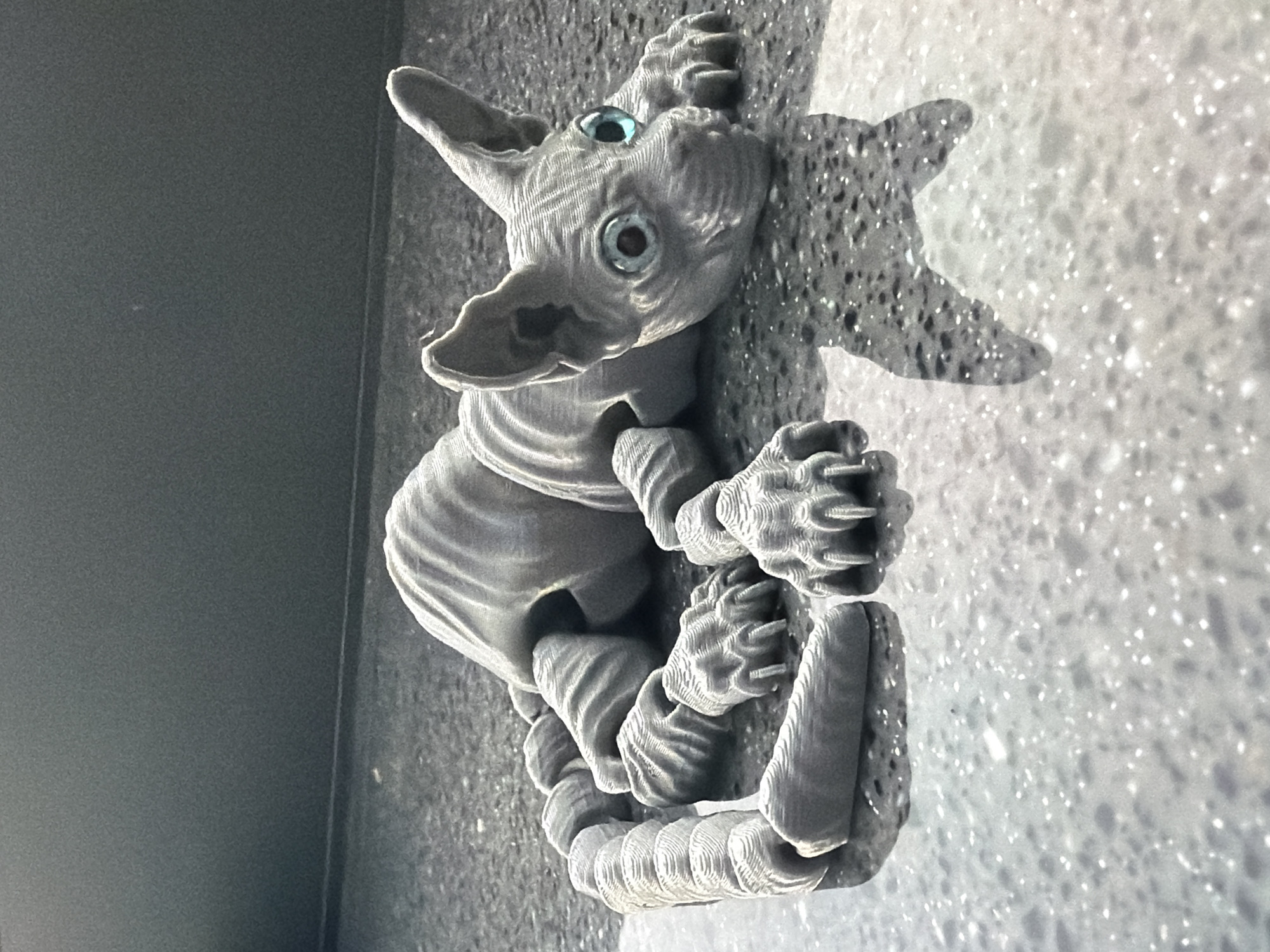 A gray, 3D-printed, segmented toy sphinx cat is lying on a reflective, speckled gray surface. The toy has large, blue-green eyes, prominent ears, and articulated paws with claws. Its body and tail are made of segments that allow for posing. The surface it rests on is wet or highly polished, creating a reflection of the toy. The background is a dark, solid, textured surface. There is no indication of the city of Moscow, Russia, or any other location.