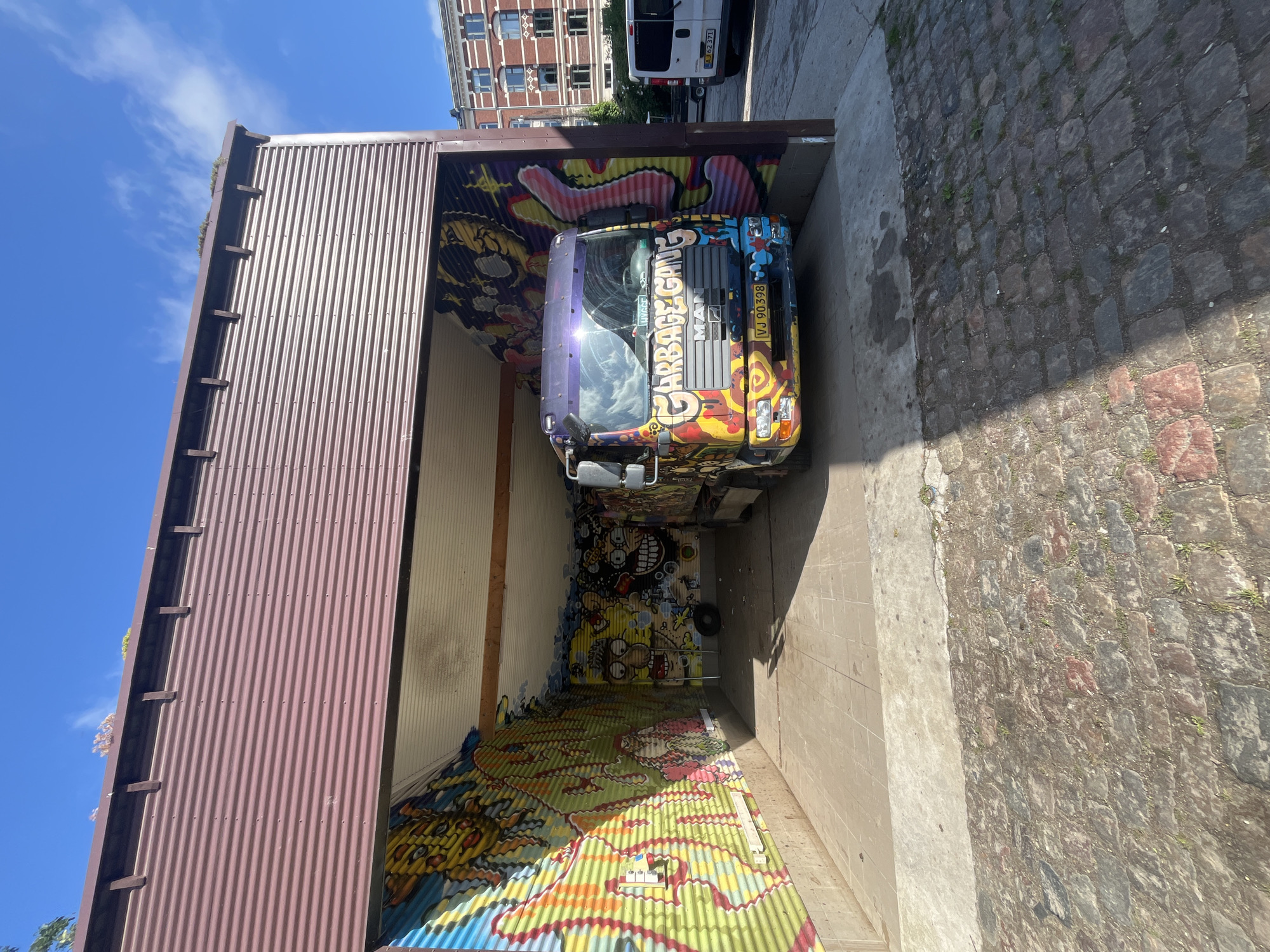 The image shows a colorful graffiti-covered garbage truck parked inside a corrugated metal structure in Copenhagen, Denmark. The structure has an open front, revealing the interior walls covered in vibrant graffiti art. The truck itself is heavily decorated with various cartoonish figures and the words "GARBAGE GANG" painted in bold letters on its front. The license plate reads "VJ 90 398". Outside the structure, a cobblestone street is visible, along with a white van parked to the right. In the background, a brick building with multiple windows suggests an urban setting. The sky is blue with some wispy clouds.