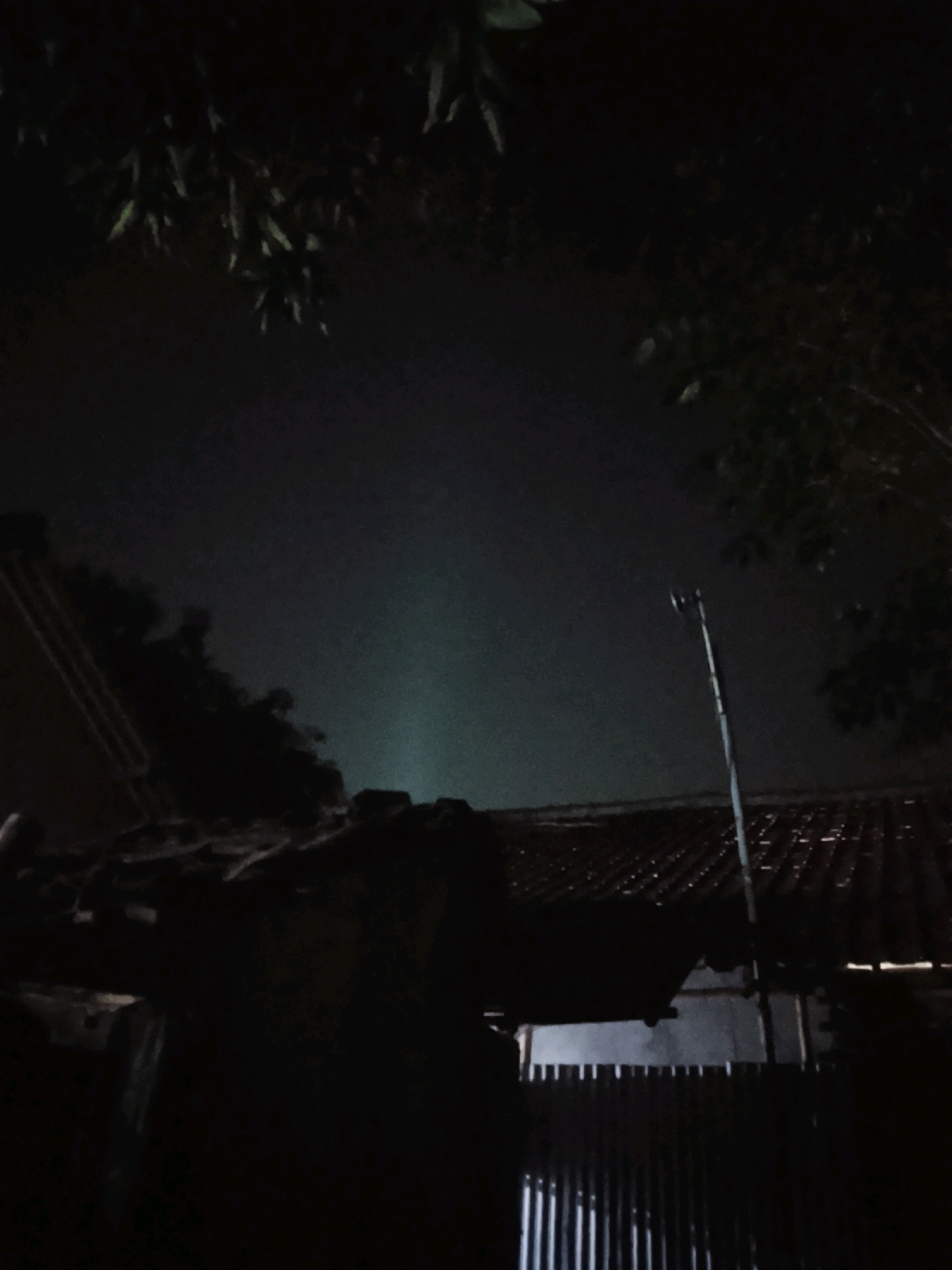 The image depicts a nighttime scene, possibly in Ujungrusi, Indonesia. In the foreground, the silhouette of a tiled roof is visible, with some debris or unevenness on its surface. A tall, slender pole, possibly a streetlight or antenna, rises vertically in the mid-ground. The background is a dark sky with a faint, greenish beam of light or fog ascending upwards. Trees with dark foliage are present in the upper portions of the frame, framing the sky and the central light beam. The overall impression is that of an outdoor, nocturnal environment.