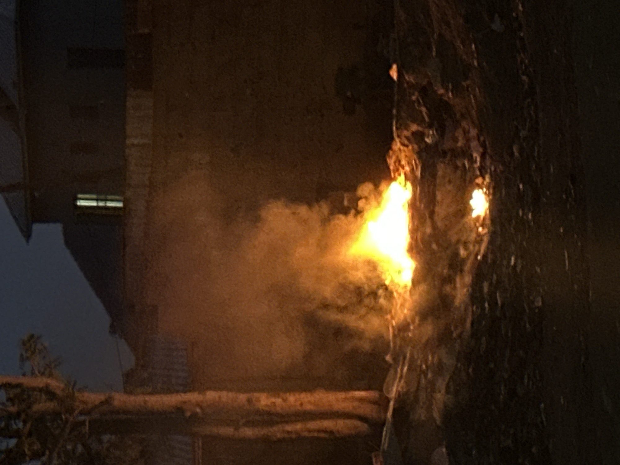 The image shows a fire burning on or near a building exterior. Smoke is emanating from the flames. A tree branch is visible in the foreground. Part of a building is seen to the left. The environment appears to be dark, possibly at night. There is no indication of the location being Bukuru, Nigeria. No people are visible.