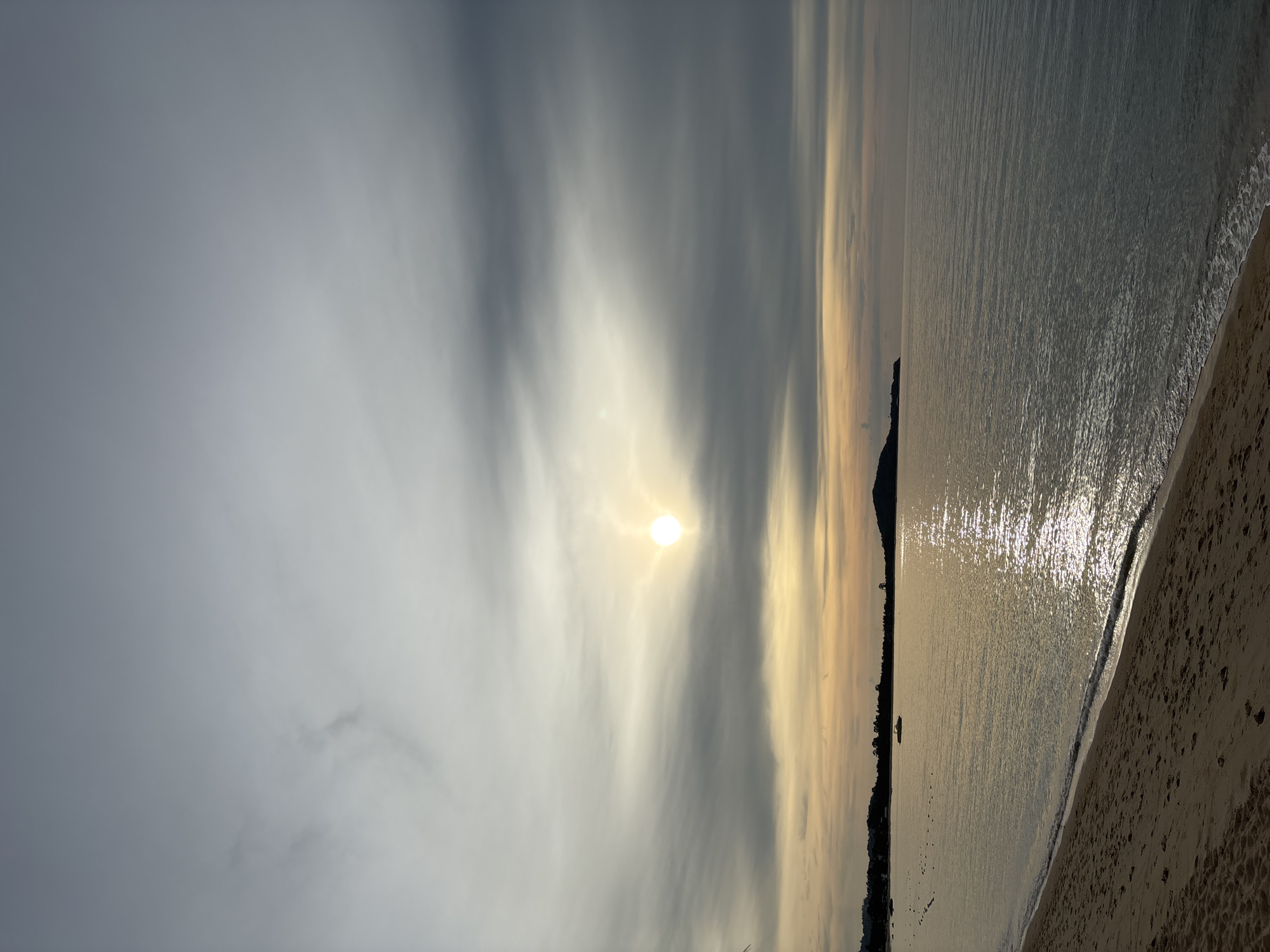 A serene beach scene in Baan Bo Phut, Thailand, captures the tranquil beauty of twilight. The sun, a bright orb nestled amongst a band of diffused clouds, casts a warm, golden glow across the sky, tingeing the horizon with hues of orange and yellow. The gentle waves of the Gulf of Thailand lap at the sandy shore, their surfaces shimmering with reflected sunlight. In the distance, a dark, mountainous silhouette marks the coastline, dotted with a smattering of trees. A small, yellow boat navigates the calm waters near the shore, its presence adding a subtle element of human activity to the otherwise untouched landscape. The sand, a soft, pale brown, shows the gentle undulation of footprints, evidence of recent visitors. The overall atmosphere is one of peaceful solitude, a perfect moment captured in time.