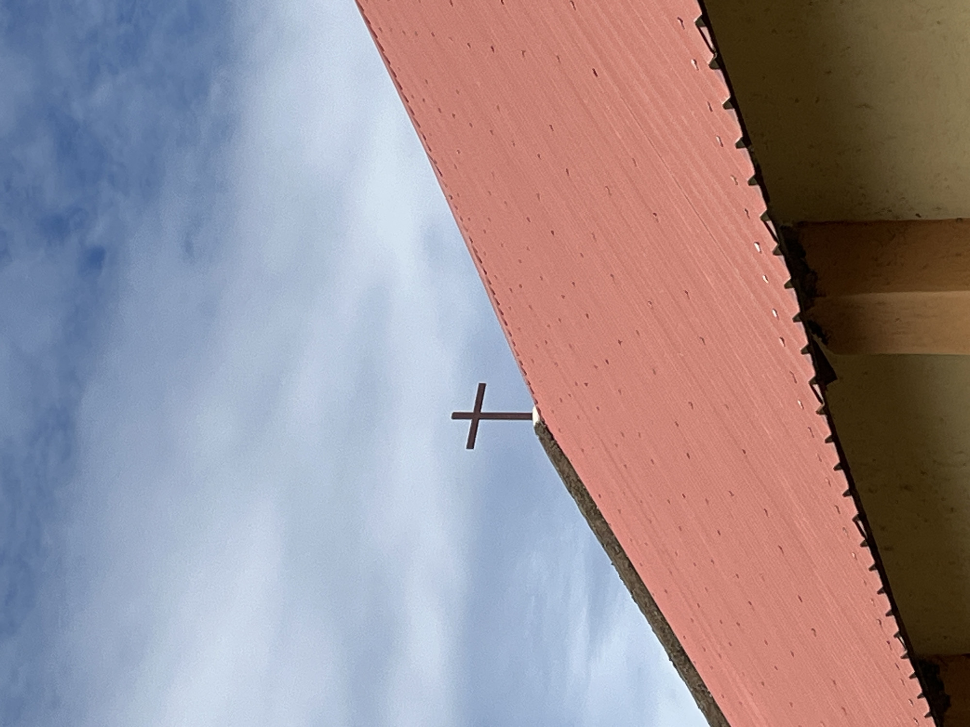 A close-up, low-angle outdoor shot captures a cross atop the red, corrugated metal roof of a building. The cross is dark brown and centrally positioned on the peak of the roof. The roof itself is angled diagonally from the bottom left to the top right of the frame, with visible fasteners dotting its surface. The building's exterior wall, made of light brown textured material, is partially visible at the bottom. Above the roof, the sky is a pale blue with scattered, wispy white clouds. The scene is static, with no people or discernible activity. The image is well-lit, suggesting daytime. The location is identified as Bawan Dodo, Nigeria.