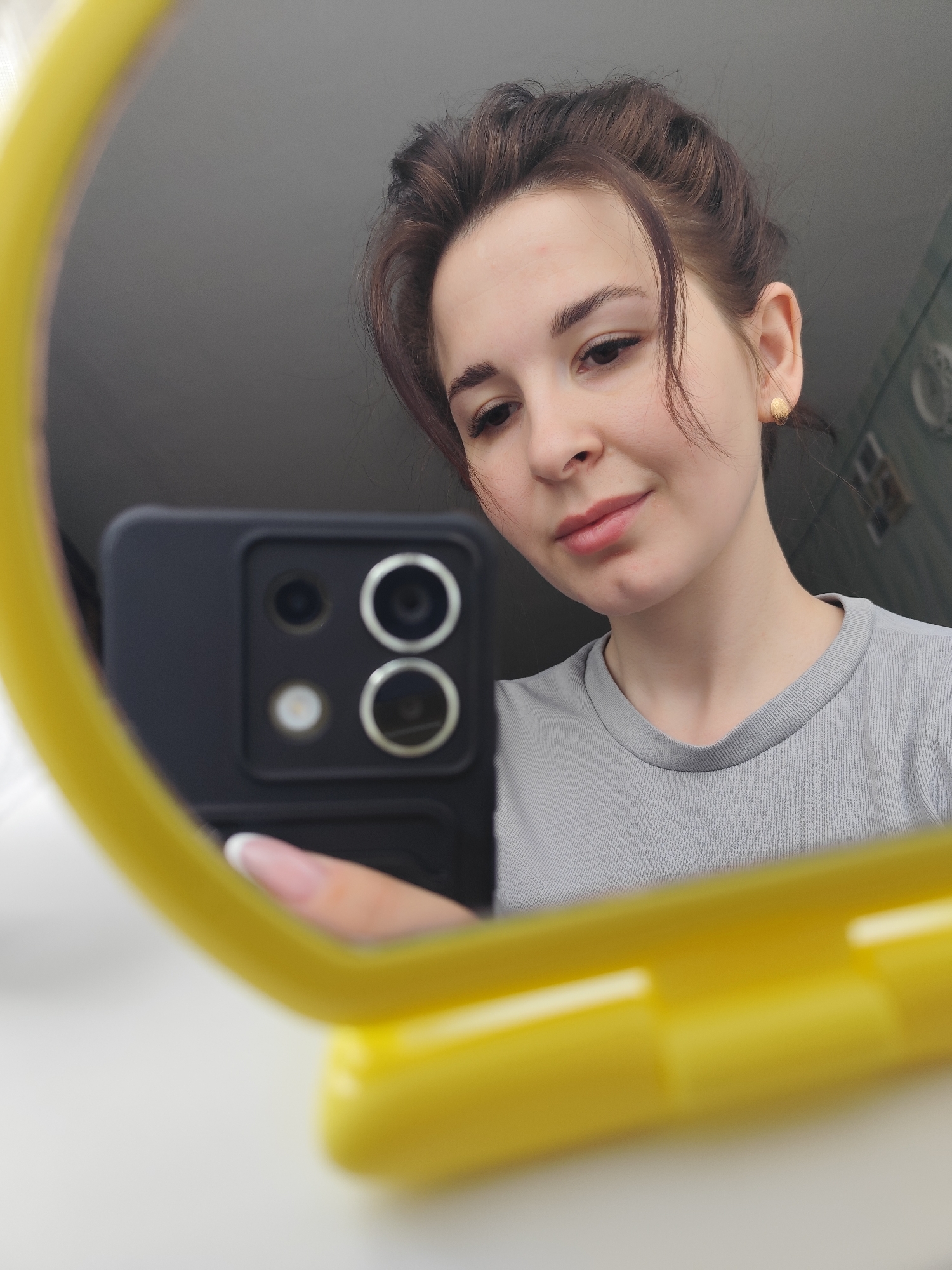 A woman is taking a selfie in a mirror. She is holding a black smartphone with a visible quad-camera setup. The mirror has a bright yellow frame. She is wearing a gray T-shirt and has her brown hair loosely pulled back. A gold stud earring is visible in her right ear. Her fingernails are manicured with a french tip style. The background is blurry. The location is not discernible from the image.