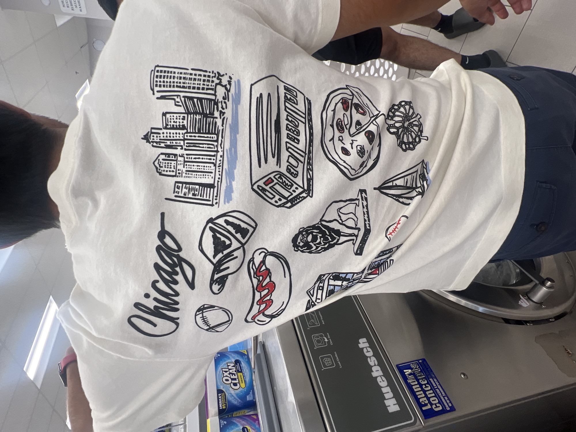Here's a concise description of the image:
The image shows the back of a person wearing a white T-shirt with a black and white graphic print referencing Chicago. The print includes iconic Chicago imagery: a skyline, a slice of pizza, hot dog, a baseball, a hat, a building, and a Chicago flag. The word "Chicago" is written in script. The person is standing in front of a Huebsch washing machine with laundry detergent nearby. The environment appears to be a laundromat. Another person is visible in the background.