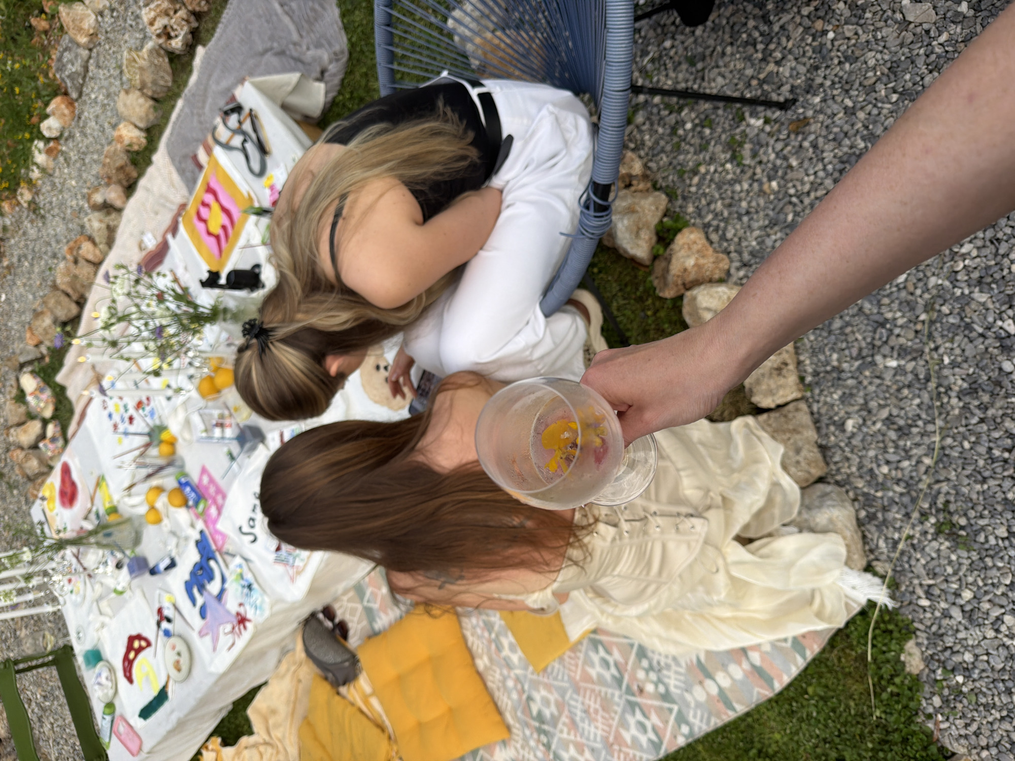 View from above shows two women sitting on a light colored blanket surrounded by rocks and gravel. A table is covered in paint, brushes and art supplies. The woman on the right is wearing a flowing light yellow dress, the woman on the left wears white pants and a black top and is seated on a blue chair. A bare arm holds a stemmed glass with flowers. Location is Dobra Voda, Montenegro.
