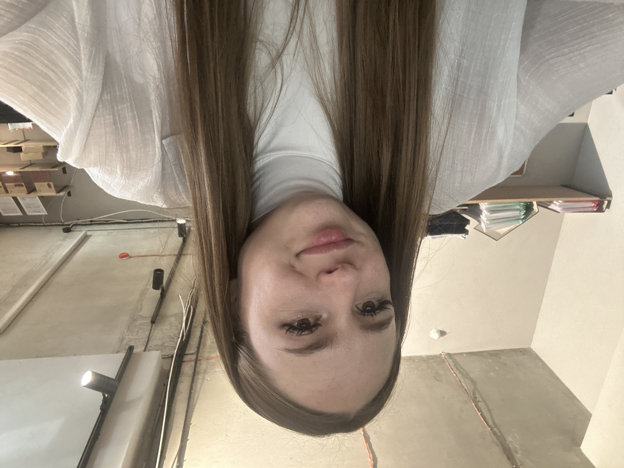 A young Caucasian woman with long, brown hair is pictured from the shoulders up, looking towards the camera with a slight smile. She is wearing a white turtleneck shirt and a white open-fronted blouse with a textured fabric. The background appears to be an indoor space, possibly an office, with a concrete ceiling, exposed wires, and industrial-style lighting fixtures. Shelves with papers and binders are visible in the background on the left and right sides of the image. The lighting suggests it is daytime, but no windows are directly visible.