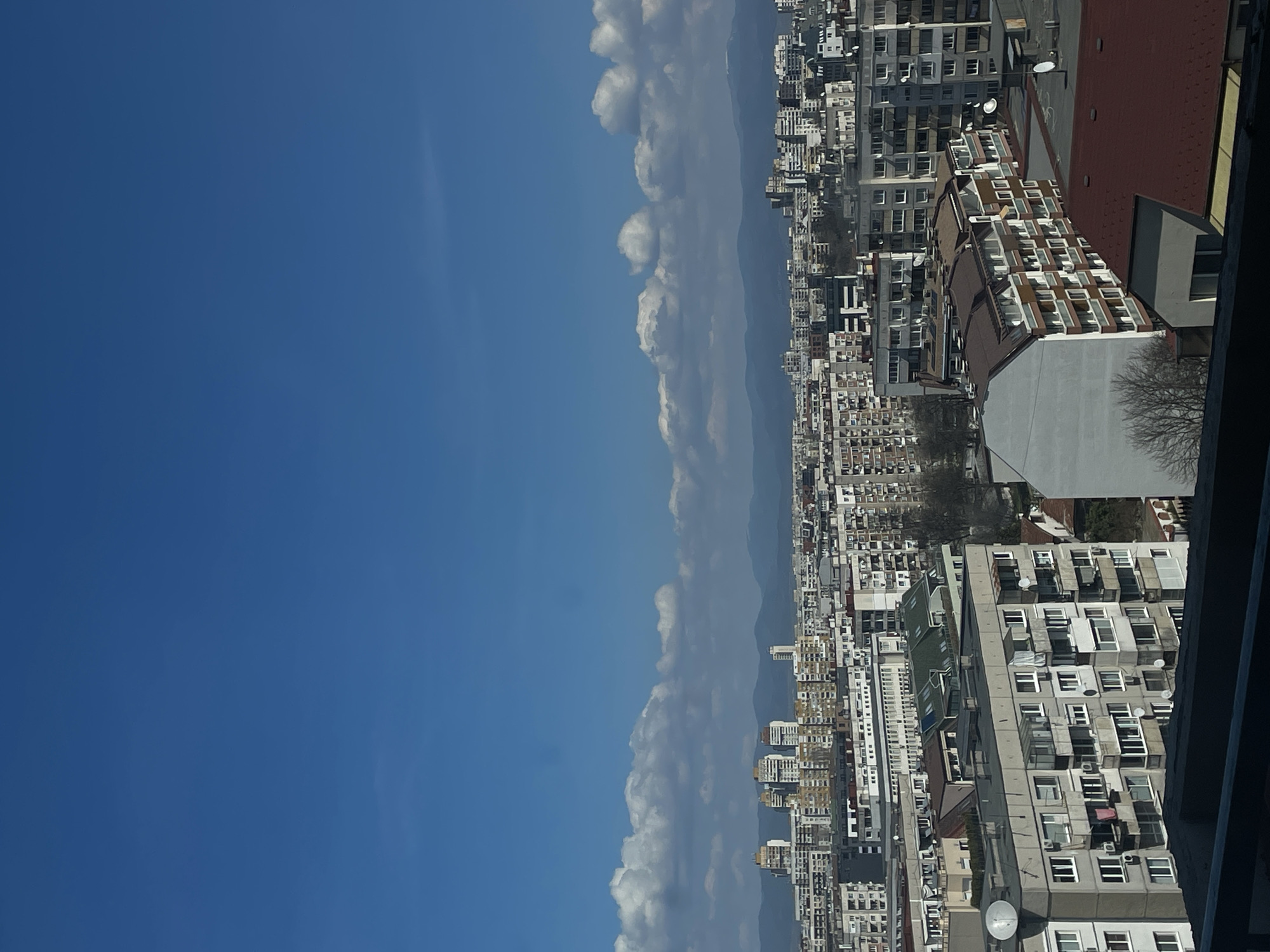 The image shows a high-angle view of a densely built urban area in Sofia, Bulgaria. The city's architecture consists primarily of residential apartment blocks of varying heights and styles, with a mix of pre- and post-socialist era construction. The buildings are predominantly white or light-colored, interspersed with some buildings having red or brown roofs. In the background, a faint outline of mountains can be seen, partially obscured by a line of fluffy white clouds. The left side of the image is dominated by a clear blue sky. There is no visible human activity. The overall environment suggests a sunny day. The perspective suggests the photograph was taken from a high vantage point, likely from another tall building or a high-rise structure.