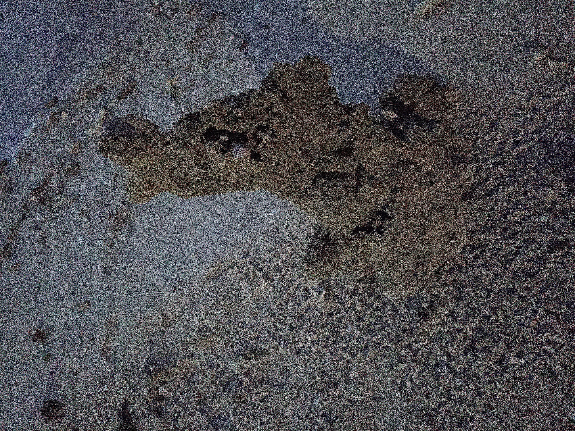 The image depicts a close-up view of a weathered and possibly eroded surface, likely stone or concrete. It is characterized by varying textures and colorations, ranging from gray to dark brown. The composition exhibits a mottled appearance with no clearly identifiable objects, people, or actions within the frame. Based on the provided location in Baan Wok Tum, Thailand, the surface could potentially be found in a natural environment, such as exposed rock formations or in a man-made environment, such as a damaged building or pavement. The image appears to be forensic in nature and suggests that the location or image is meant to be used as a study for a crime scene.