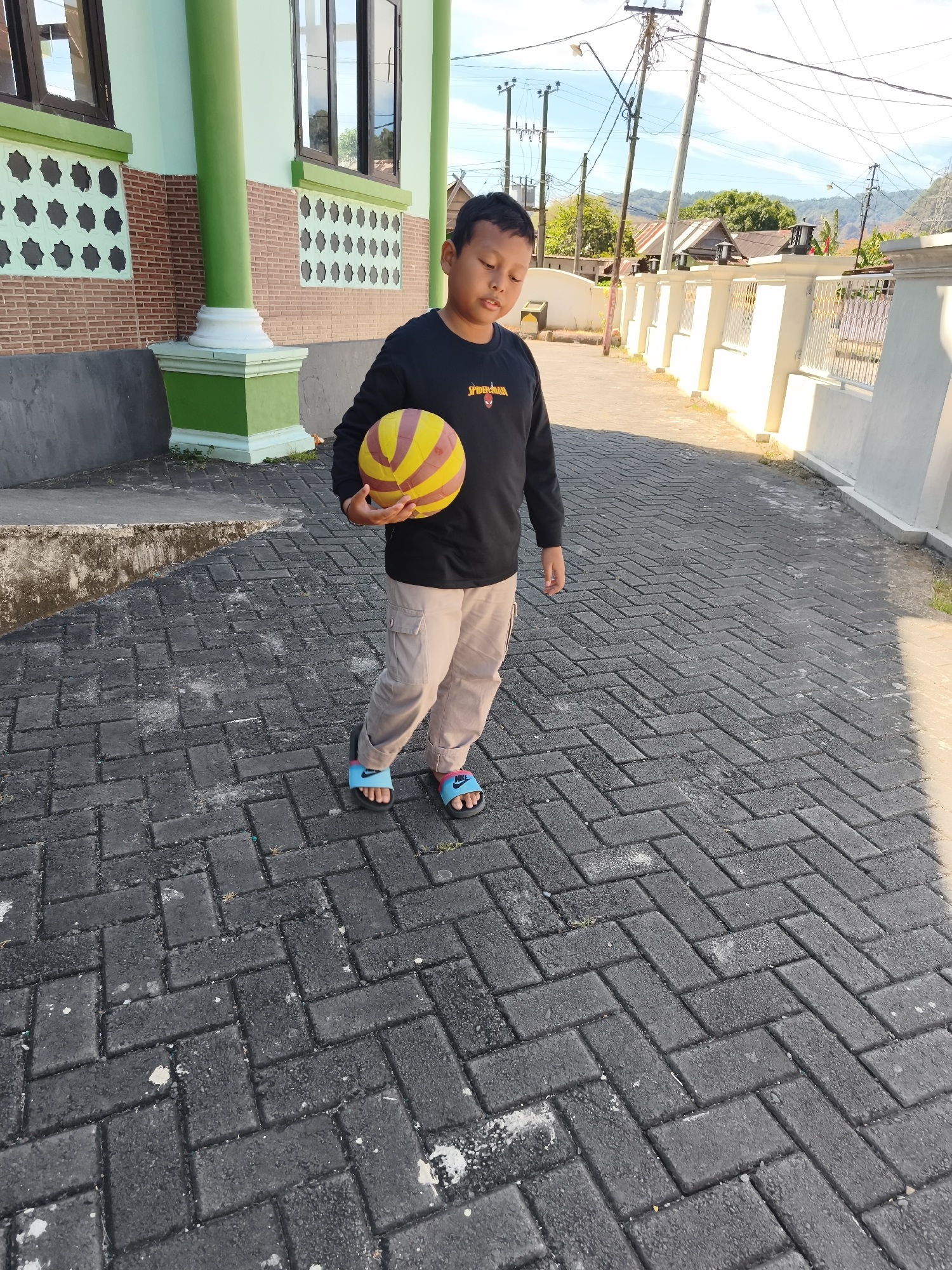 A young boy stands on a brick-paved walkway, holding a yellow and red ball. He is wearing a black long-sleeved shirt with a "Spider-Man" graphic and beige cargo pants. The boy is looking down, with his eyes closed. He is wearing blue and pink slide sandals. Behind him, there is a building with green pillars and a brick facade, and further in the background, there are houses, power lines, and trees under a bright blue sky. The scene appears to be in Tonasa, Indonesia.