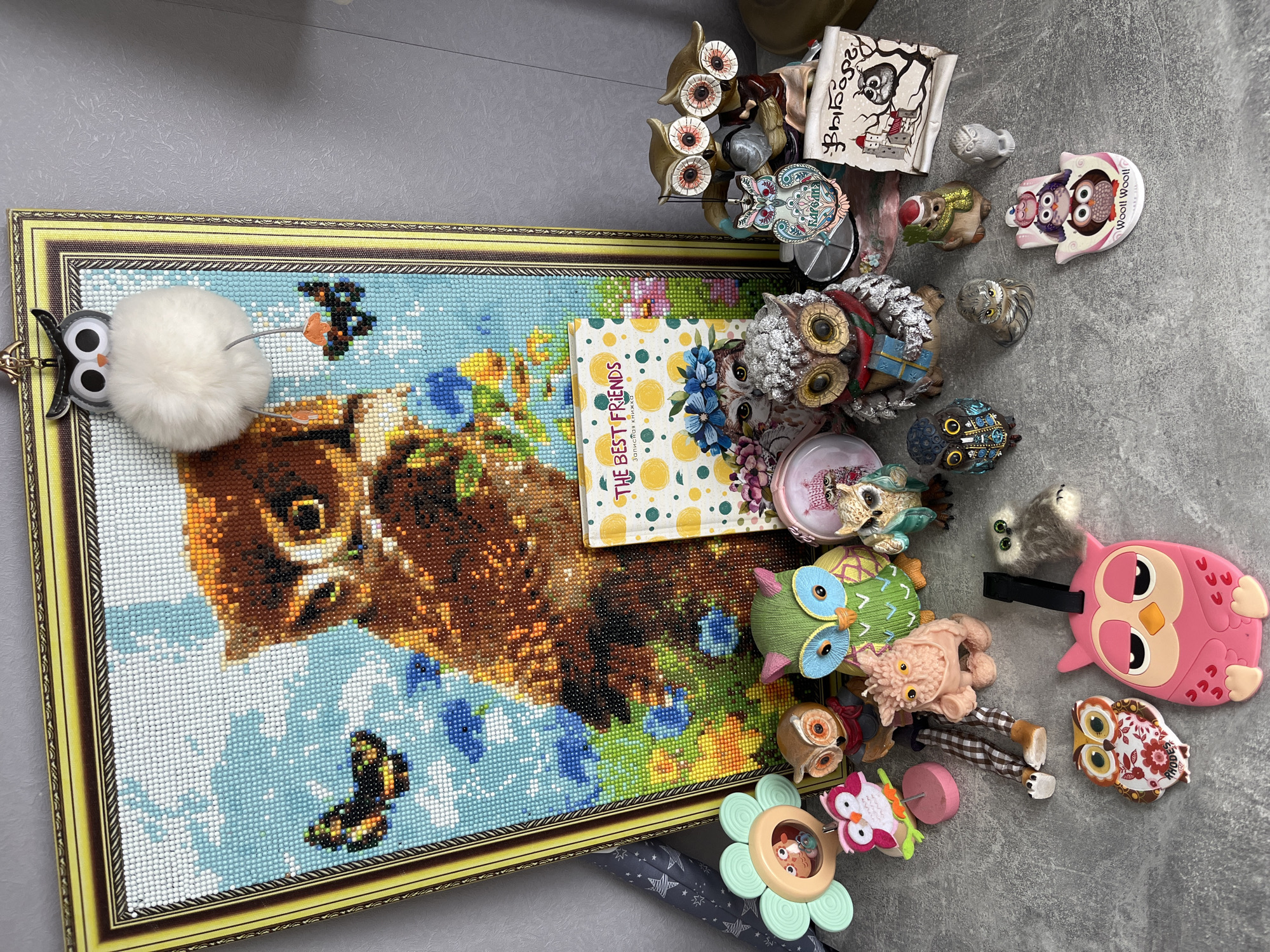 The image shows a collection of owl-themed items arranged on a gray surface, likely a floor or table. Dominating the scene is a framed picture of an owl, created with a diamond art technique. Atop the picture lies a yellow notebook with "THE BEST FRIENDS" written on the cover in English and Russian, along with a polka dot design. Surrounding the picture are numerous owl figurines, toys, and decorations of various materials and styles: ceramic, plush, plastic, and metal. A few items are potentially functioning objects like a flower-shaped toy with an owl inside. The items are closely grouped, with some objects overlapping. The overall impression is a collection of owl-related items. The word "Vyborg" is visible, which is a city in Russia. The picture also contains the word "PHODES". This image was taken in the city of Shushary, Russia.