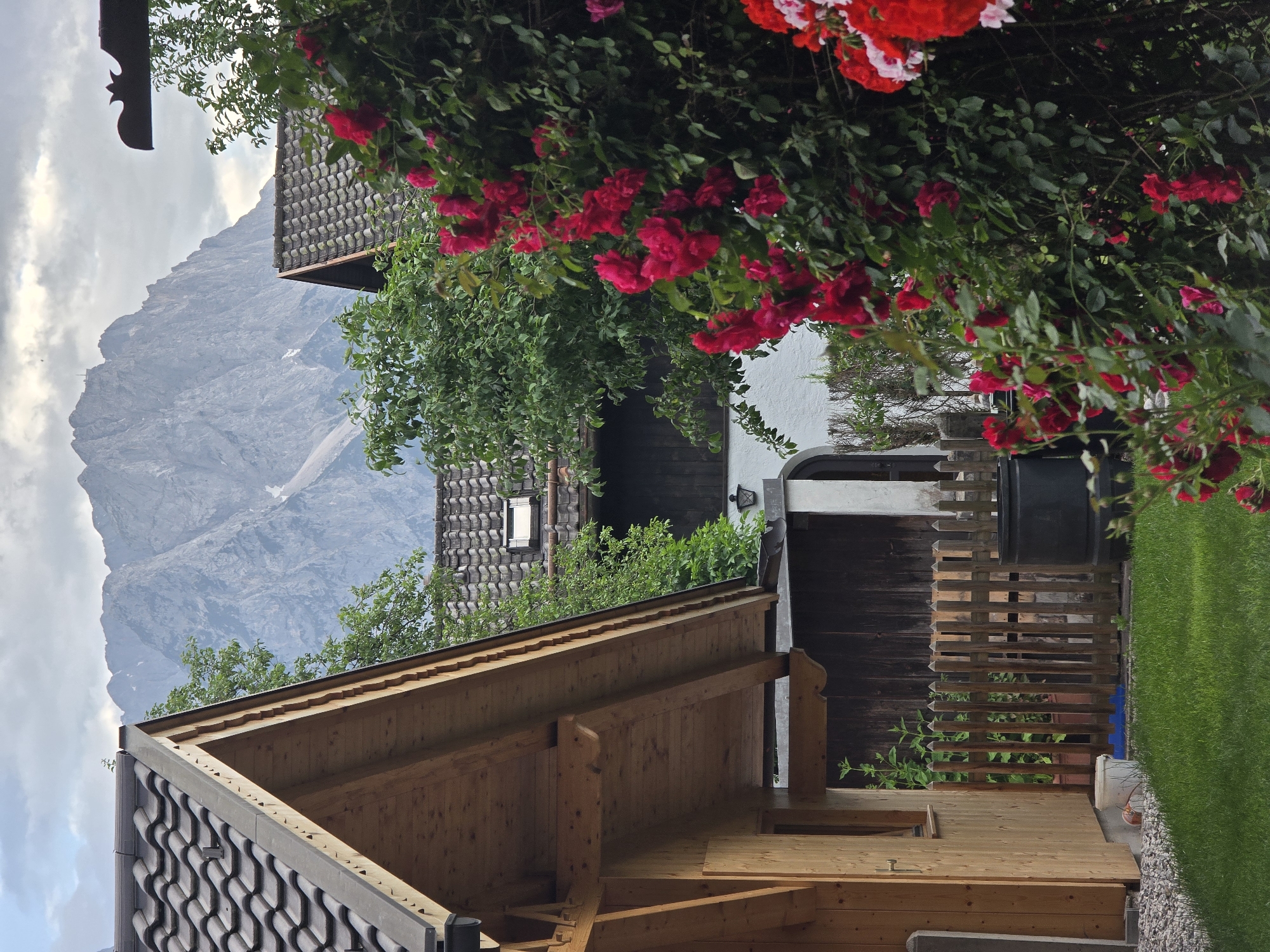 This image depicts a residential scene in Garmisch-Partenkirchen, Germany, with a prominent mountain range in the background. In the foreground, a wooden structure, likely a house or shed with a tiled roof, is visible on the left. Adjacent to it is a lush green lawn and a fence made of vertical wooden planks. A large bush with vibrant red roses is in bloom on the right side of the frame, partially obscuring another building with a similar tiled roof. The mountainous backdrop is imposing, with rugged peaks and patches of snow, suggesting a high-altitude location. The sky is partly cloudy, with a soft, diffused light. The overall impression is one of a tranquil, rural setting within a scenic mountainous region. There are no people visible in the image.