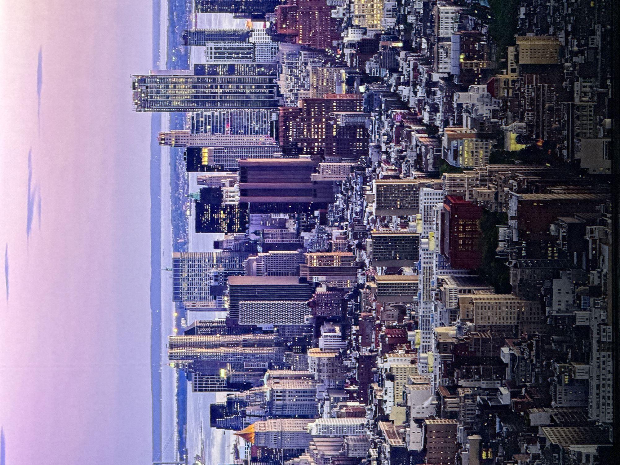 The image is a wide aerial shot of a densely packed cityscape dominated by numerous skyscrapers of varying heights and architectural styles. The sky above is a soft gradient of purple and pink, indicating either sunrise or sunset. The buildings are illuminated, with many windows showing lights, suggesting it is dusk or evening. In the distance, a body of water is visible, with landmasses on the horizon and what appear to be bridges. The architecture suggests a major metropolitan area, likely in North America, given the style of the buildings. The cityscape is intricate and detailed, with a mix of older and modern structures. The image does not depict any people. The lighting and perspective create a sense of scale and grandeur. There is no indication of the city being Saratov, Russia.