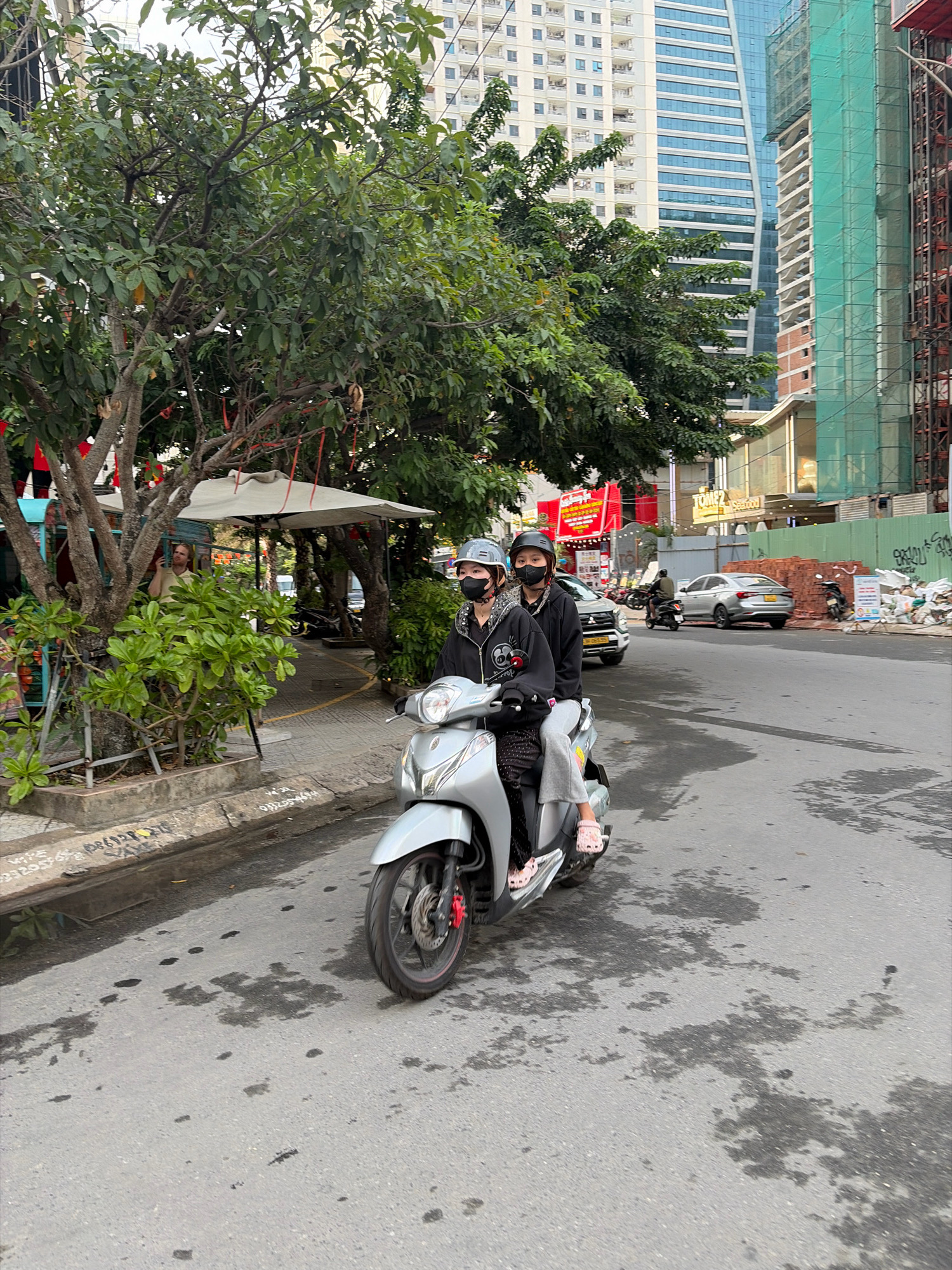 A scooter with two passengers drives on a city street in Đà Nẵng, Vietnam, with a modern building and construction site in the background. The driver and passenger are wearing helmets and face masks. The scooter is silver and the road surface shows oil stains. On the left, there are trees and a sidewalk cafe.