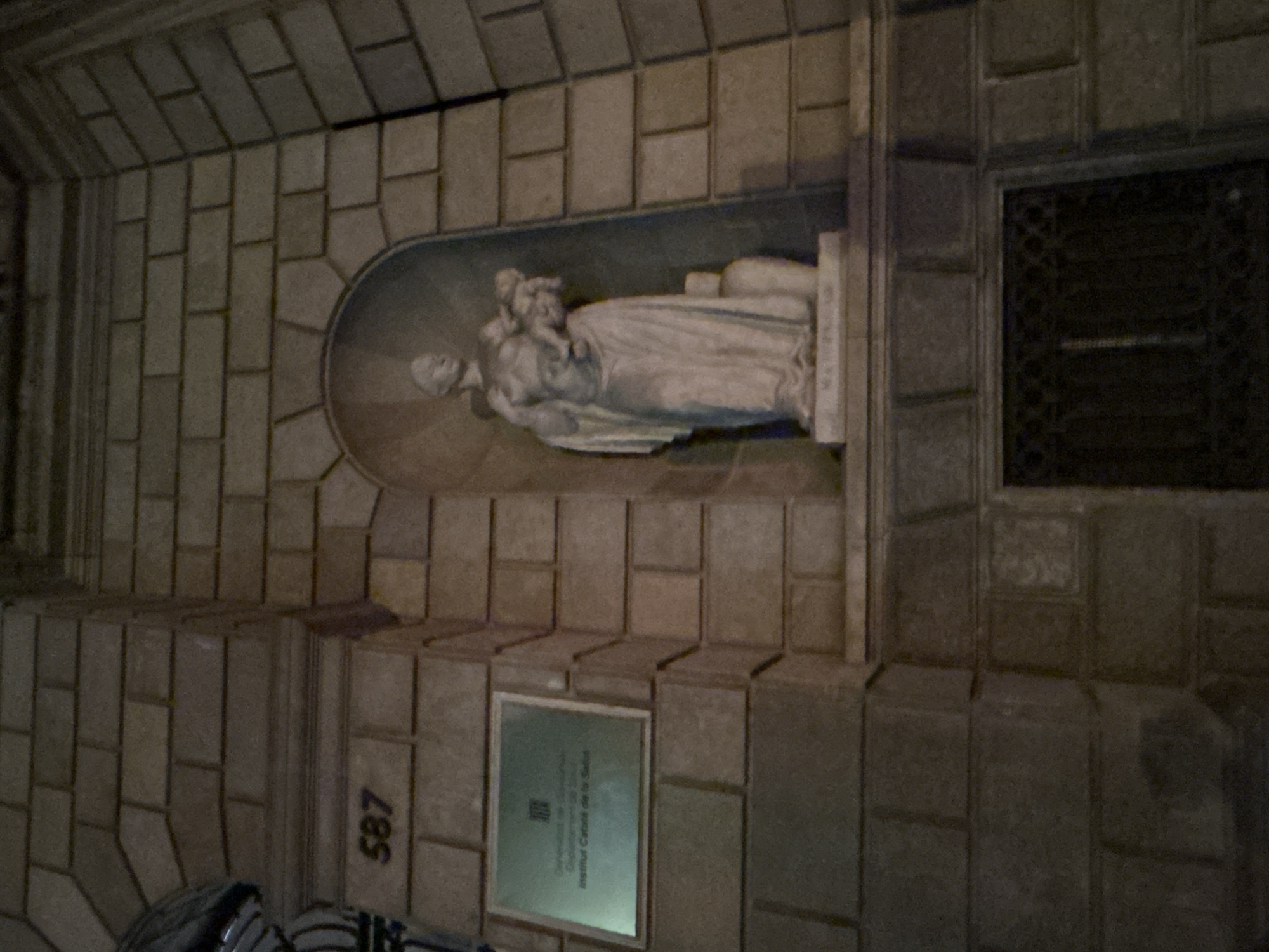 The photograph shows an exterior wall in Barcelona, Spain. The wall is constructed of grey brick and contains a stone sculpture depicting a woman holding a child, set within an arched niche. Below the sculpture is a rectangular plaque mounted on the wall. To the right of the sculpture is a dark, square opening with an ornate metal grate. The number "587" is visible on the wall near the bottom left corner. The lighting is dim, suggesting it might be evening or a shaded area. The scene is static and appears to be a historical or architectural detail of a building facade.