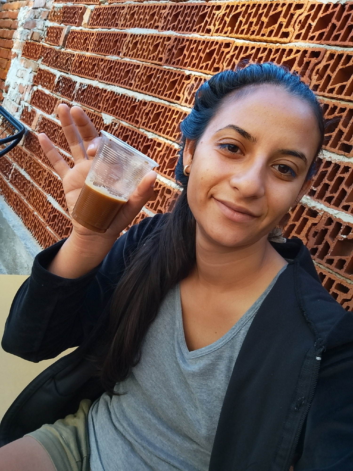 A young woman with dark hair pulled back is holding up a clear plastic cup filled with a brown liquid, likely coffee, in front of a textured brick wall. She is smiling at the camera. She is wearing a black jacket over a gray t-shirt. The background is a wall made of reddish-brown bricks with visible holes. The scene appears to be outdoors, possibly on a sunny day, given the lighting. The location is Yambol, Bulgaria.