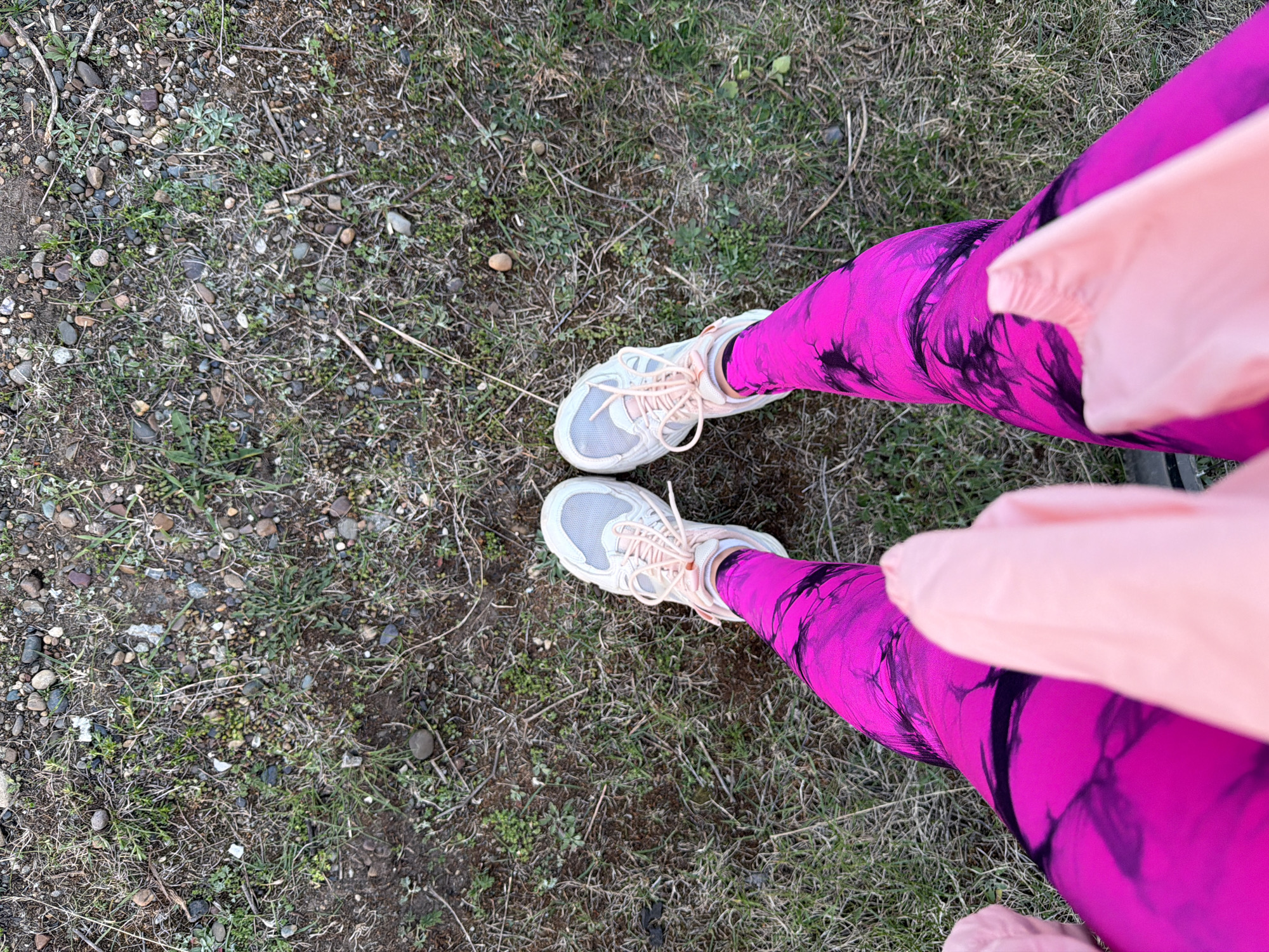 The image shows a person standing on a patch of ground in what appears to be an outdoor area. The ground is a mix of dirt, grass, small rocks, and dry vegetation. The person is wearing bright pink and black tie-dye leggings and light gray running shoes with light pink laces. A light pink garment or accessory is draped over the right leg. The location is unknown and cannot be determined from the image. There is no discernible information in the image indicating the city of Chaikovsky, Russia.