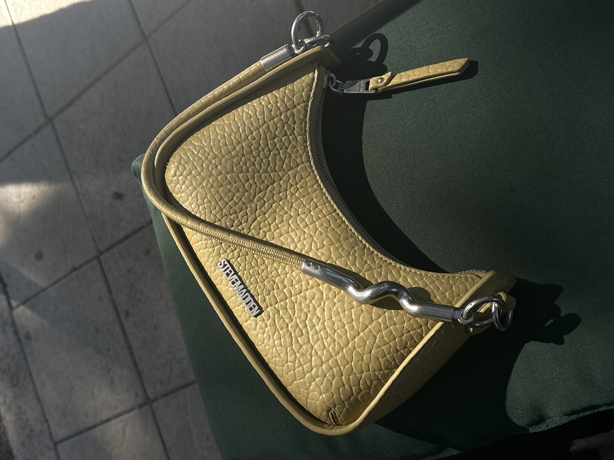A yellow textured handbag with a silver Steve Madden logo is placed on a dark green surface. The handbag has a zipper closure and a single strap with a silver chain detail. The background shows a tiled surface, possibly a sidewalk, with shadows from sunlight. The scene is outdoors.