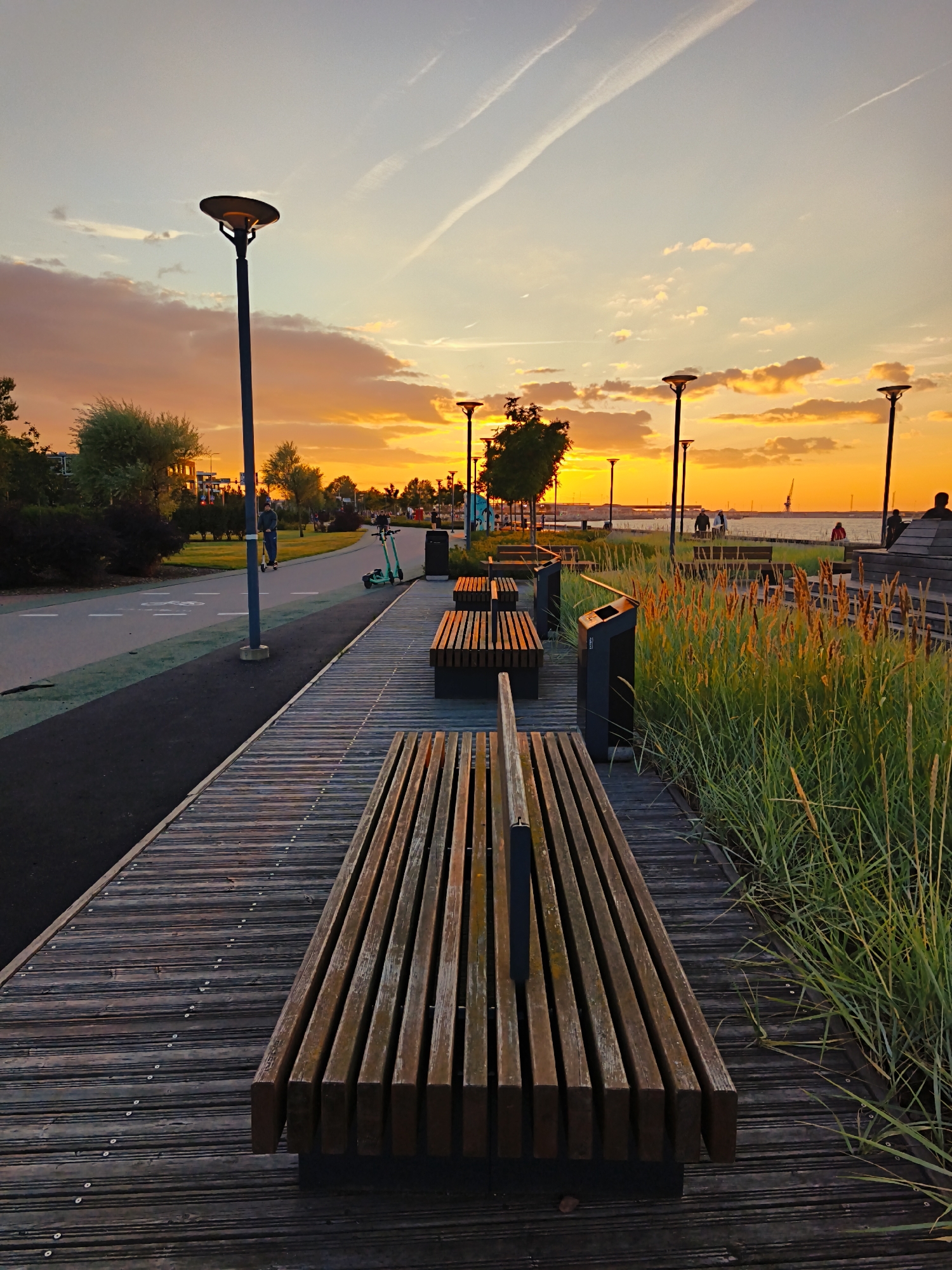 This image captures a sunset scene at a waterfront promenade in Tallinn, Estonia. The sky is ablaze with warm orange and yellow hues, with streaks of contrails visible against a pale blue backdrop. In the foreground, a wooden boardwalk stretches into the distance, lined with modern wooden benches and strategically placed trash receptacles. To the left, a paved path curves alongside a grassy area and rows of trees, with streetlights illuminating the walkway. A person is seen riding an electric scooter on this path, while another scooter is parked nearby. Further down the boardwalk, several more benches are visible, some occupied by silhouetted figures. Beyond the promenade, a body of water, likely the Baltic Sea, is calm, with a few distant boats and industrial structures on the horizon. Lush green grasses and reeds flank the boardwalk, adding a natural element to the urban setting. The overall atmosphere is one of peaceful evening recreation.