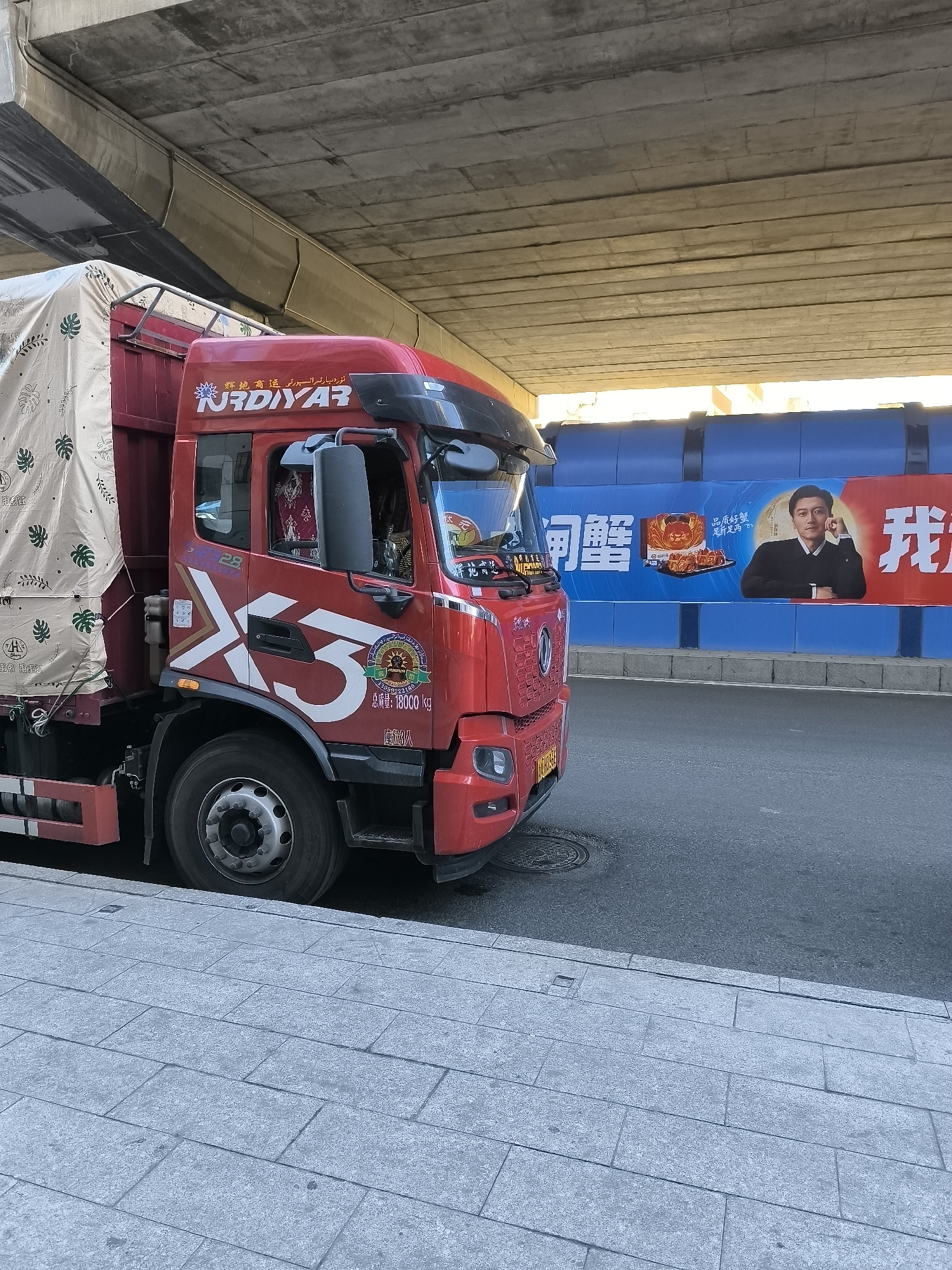 The image depicts a red truck parked on the side of a road under an overpass in Ürümqi, China. The truck has a white "X3" logo on its side. A large banner hangs on a blue barrier behind the truck. The banner features Chinese text, an image of crabs, and a portrait of a man. The overpass is constructed of concrete. The sidewalk is paved with grey rectangular stones. The scene appears to be an urban environment with road infrastructure and advertising.