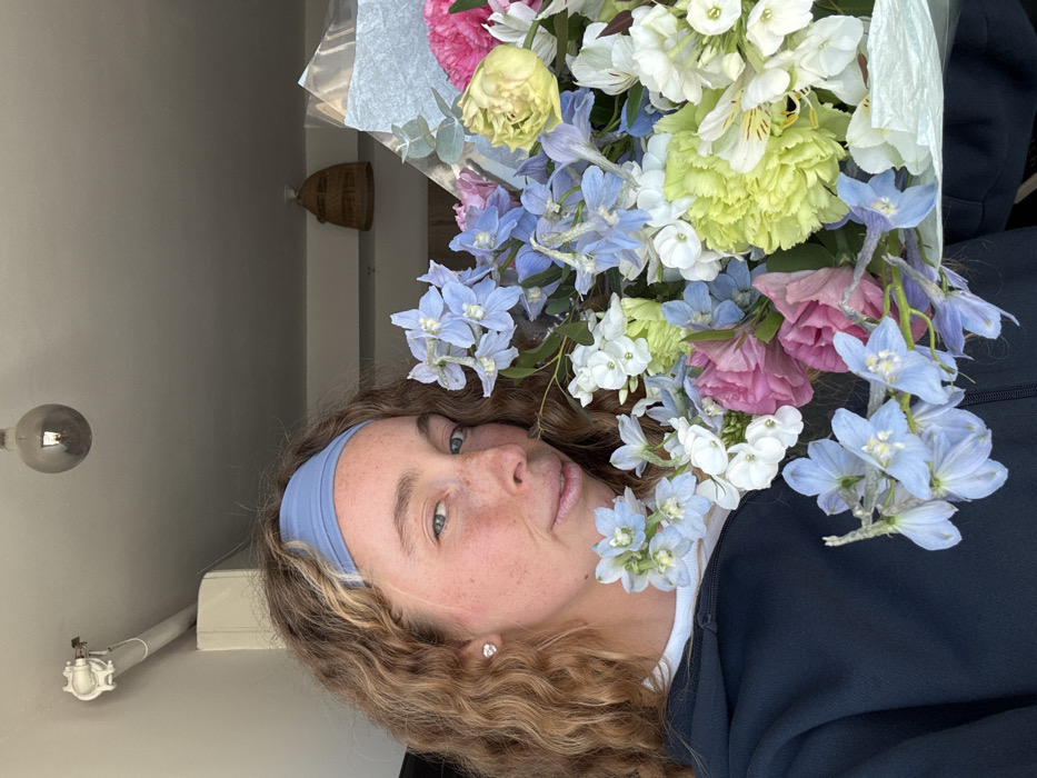 A female subject, appearing to be in her mid-20s to early 30s, is captured in a frontal, chest-up portrait within an indoor setting in the city of Tel-Aviv, Israel. She has fair skin with visible freckles across her nose and cheeks, light-colored eyes, and shoulder-length, curly auburn hair, secured by a light blue fabric headband. A small, clear or silver stud earring is visible in her left ear. She wears a dark blue or black hooded sweatshirt over a white inner layer. Her lips exhibit a glossy texture, and she displays a slight smile, engaging directly with the camera.
She is holding a large, vibrantly colored floral bouquet that dominates the right half of the frame. The bouquet contains a variety of flowers, including numerous light blue delphiniums or larkspurs, clusters of small white flowers, ruffled light green carnations, pink or mauve eustomas, and white alstroemerias, interspersed with green foliage. The bouquet is wrapped in a crinkled, translucent light blue or silver-toned paper or film.
The background consists of a light-colored ceiling. An exposed, smoked-glass light bulb hangs from the ceiling in the upper-left quadrant. To the upper-left, a white wall-mounted fixture with a curved pipe, possibly an air conditioning component or electrical conduit, is visible. Another decorative, woven straw or rattan basket-like object hangs from the ceiling in the upper-right. The lighting appears bright and evenly distributed.