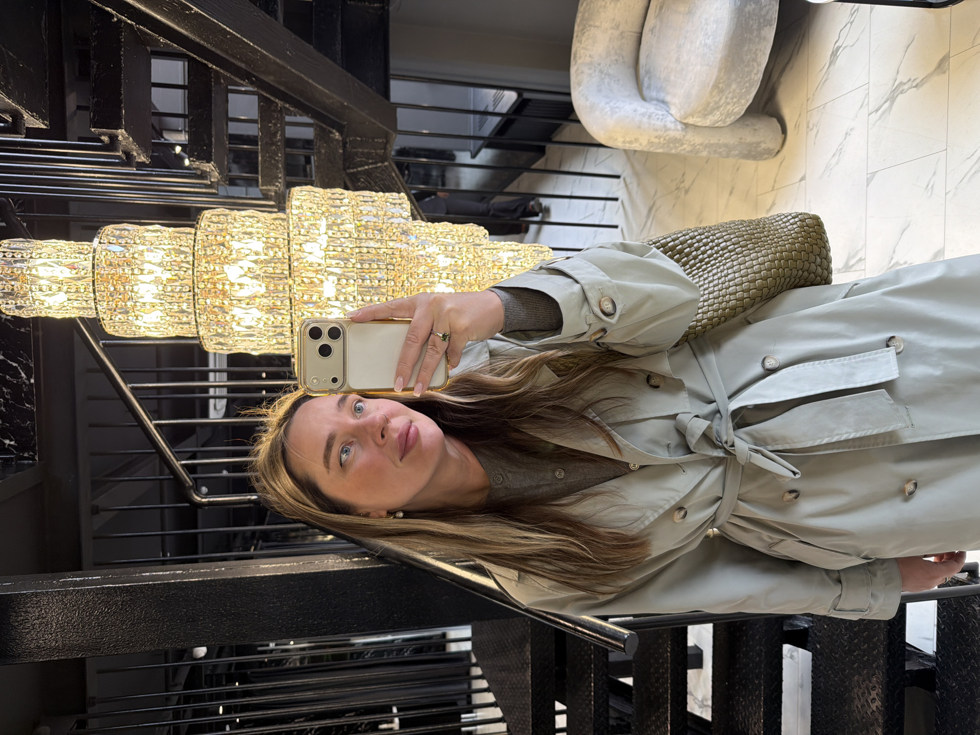 A woman takes a selfie in front of a multi-tiered chandelier and black metal staircase with horizontal bars, appearing to be indoors. She wears a light-colored trench coat tied at the waist, and holds a smartphone in her right hand. A wicker purse is visible. Behind her, there is a white marble floor and a curved chair. The scene is at the Radisson Collection Hotel Moscow, Russia.