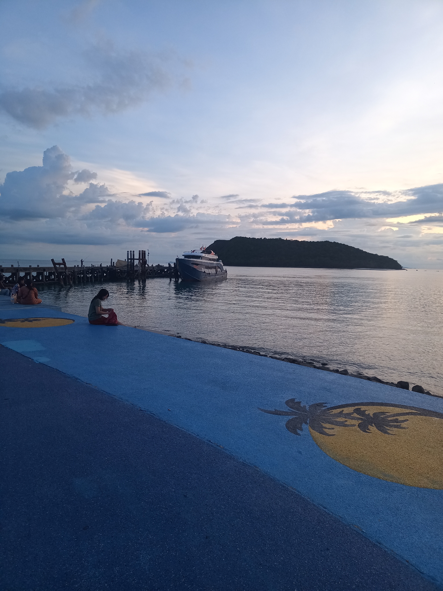The image depicts a waterfront scene at dusk on Koh Phangan, Thailand. In the foreground, a blue pathway with a yellow circular design featuring a palm tree motif runs along the edge of the water. A person is seated on this pathway, facing the water, with their legs bent and wearing a green shirt and red skirt. To the left, another person is seated on the same pathway. Beyond the pathway, a body of water reflects the dimming sky. A wooden pier extends into the water, with a modern ferry docked beside it. The ferry is dark gray with white accents and a red stripe on its bow. The water is calm, and a distant island with lush green vegetation is visible on the horizon. The sky above is a gradient of pale blue and white, with scattered clouds. The overall atmosphere is serene and tranquil.