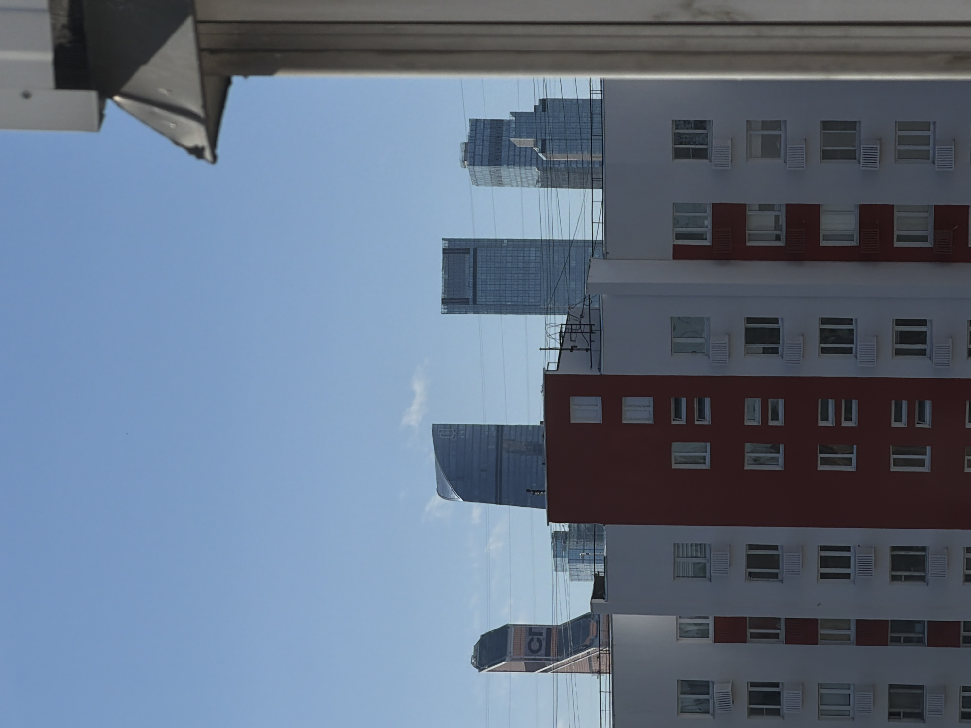 The image shows a view from a building in Khimki, Russia. In the background are several modern skyscrapers. In the foreground is another building with white and red sections and many windows. The sky is clear and blue. There are some utility lines visible in the space between the buildings. A small portion of the building the photo was taken from is visible at the top left of the frame.