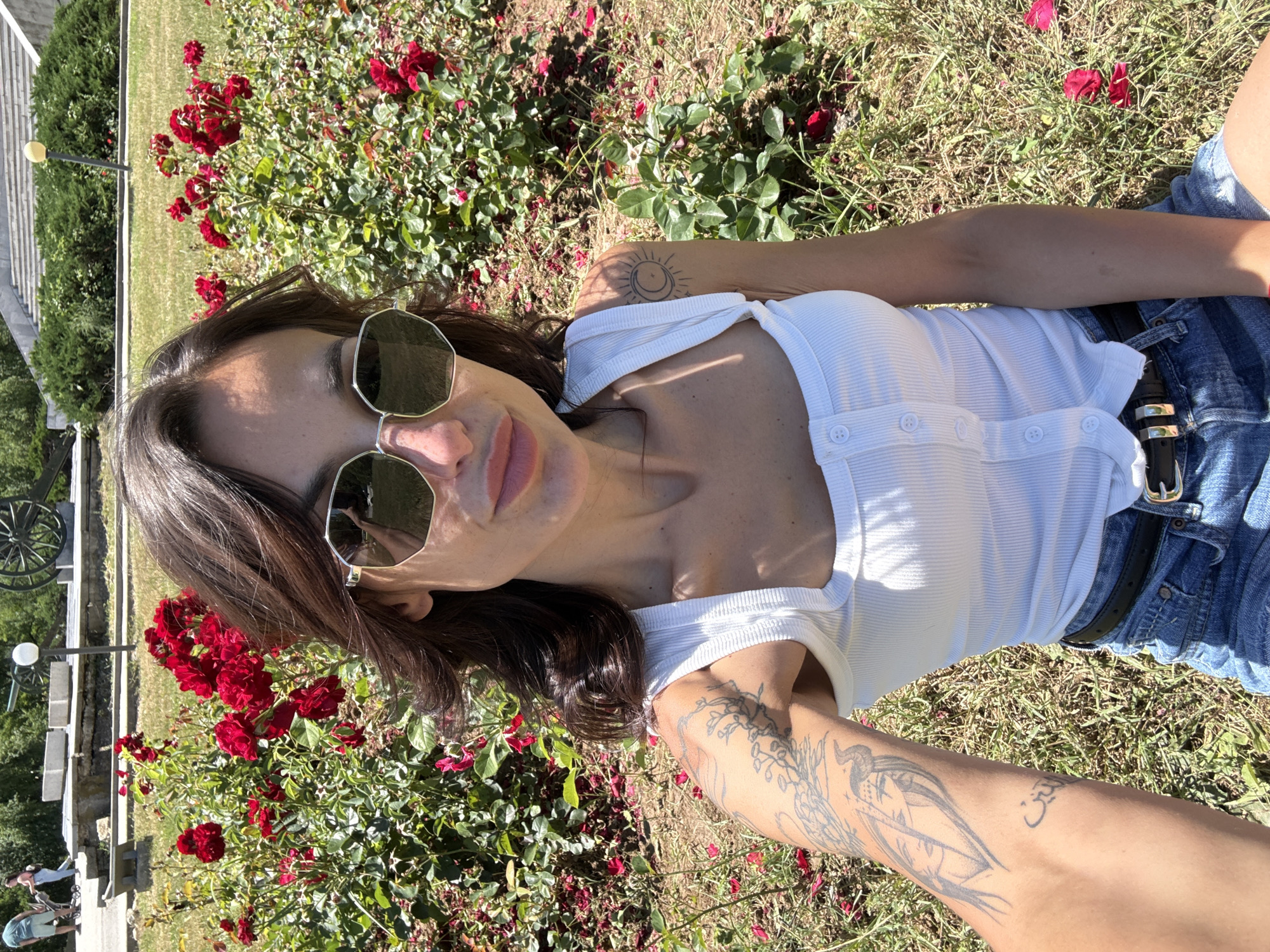 A woman is posing for a selfie in a park with red roses in Pleven, Bulgaria. She is wearing a white tank top, denim shorts, and sunglasses. Her arm is extended towards the camera, revealing tattoos. The background shows rose bushes, grass, and a monument with cannons.