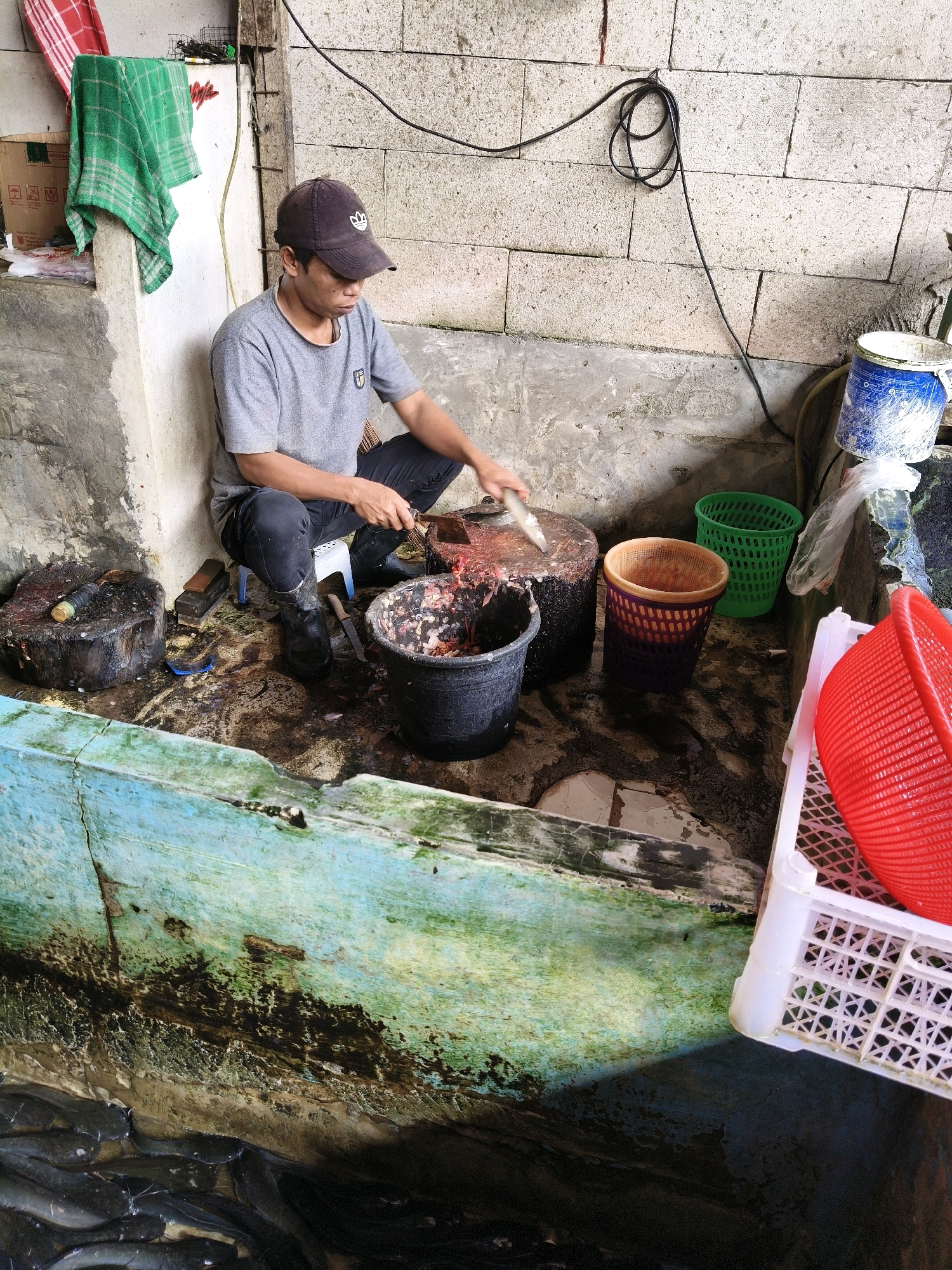 A man wearing a grey t-shirt and black pants, along with black boots and a dark baseball cap, is shown squatting and preparing fish in an outdoor setting. He is seated on a small white stool. In front of him, a large round chopping block is positioned on the ground, and he is actively using a knife to cut a fish. To his right, a black bucket filled with fish parts and what appears to be blood is situated. Nearby, there are several baskets: a green one, a purple and orange one, and a red one placed inside a white plastic crate. The environment is gritty and appears to be a market or processing area, with concrete walls and flooring that show signs of wear and dampness. A trough filled with live fish is visible in the foreground. The scene is lit by overhead light, casting shadows. The location is identified as Setiamekar, Indonesia.