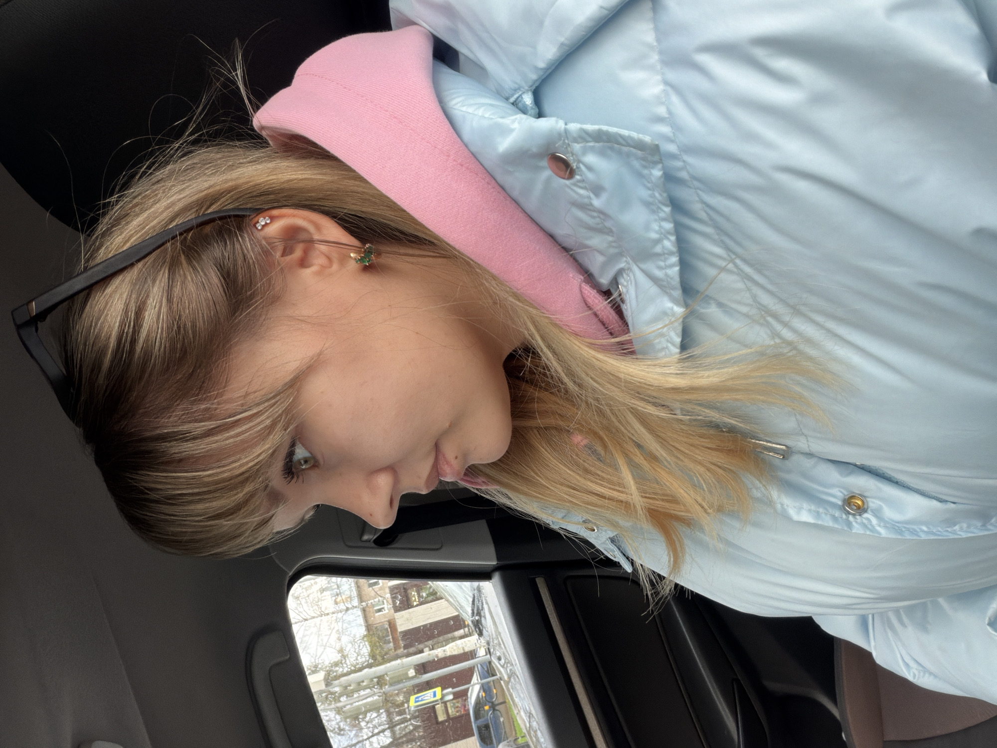 A young woman is captured in a profile shot inside a vehicle, with the image rotated counter-clockwise. She has blonde or light brown hair and is looking downwards and slightly away from the camera. Dark-framed sunglasses are perched on her head or hair. Her right ear is adorned with multiple earrings: three small white stones arranged in a cluster higher on the ear, and a small, green, possibly floral-shaped earring in the lobe.
She is wearing a light blue puffer jacket layered over a pink hooded sweatshirt, with the pink hood visible around her neck. The puffer jacket has a slightly reflective texture and features metallic snap buttons.
The background reveals the dark interior of what appears to be a car, including portions of the headliner. Through the car window, a street scene is visible, featuring buildings, bare trees suggesting a colder season like late autumn or winter, and a yellow road sign. The lighting indicates it is daytime. Her expression is neutral, appearing contemplative or focused on something out of view below her. No discernible text is visible in the image.
