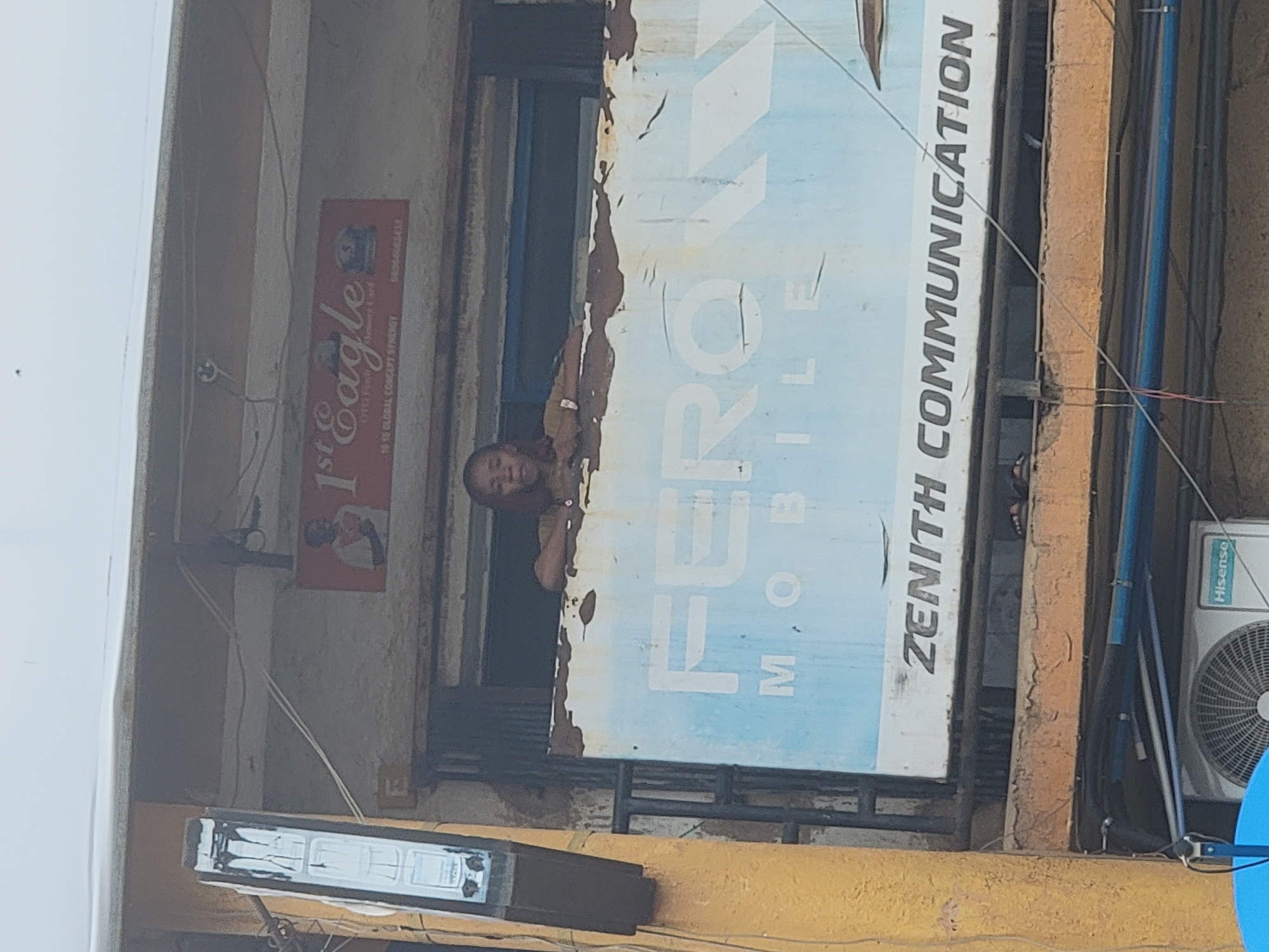 A medium shot captures a building with signs and a person, possibly in Jos, Nigeria. The image is rotated, presenting a vertical perspective. At the center, a man peers from behind a large, rectangular sign that reads "FEROZ MOBILE ZENITH COMMUNICATION." The sign is weathered, exhibiting peeling paint and rust. To the left of the Zenith Communication sign, a smaller sign for "1st Eagle" is visible, also weathered and slightly obscured. Electrical wires are strung across the building's facade. To the right, a Hisense air conditioning unit is mounted. The building itself appears to be aged and in need of repair. The color palette is dominated by faded hues of blue, white, red, and brown.