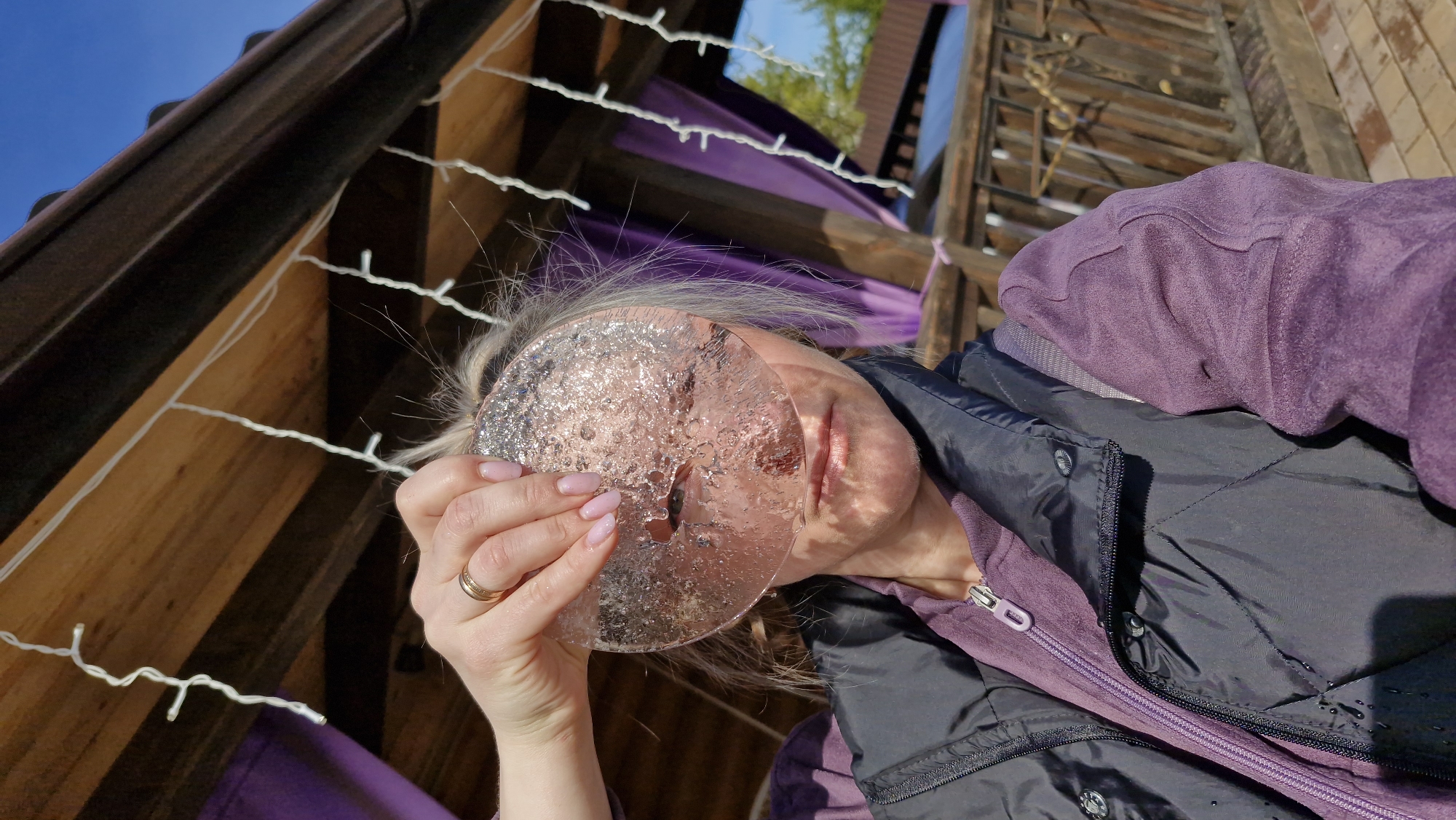 A woman with fair skin and light blonde hair is holding a thin, clear, circular ice disc in front of her face. She is wearing a purple hoodie underneath a black, quilted jacket. She has light pink nail polish on her fingernails and a gold wedding band on her ring finger. Her face is partially visible through the ice, distorted by the frozen texture.
The location appears to be outdoors, beneath a wooden structure possibly associated with an outdoor space or building. White string lights are hung across the structure. Purple fabric is draped in the background. The sky is visible above, with hints of blue sky and some green foliage, indicating an open-air environment. The image is angled upwards, capturing the woman's face and the surrounding structure. There are no explicit markers indicating this is specifically Otchor, Russia.