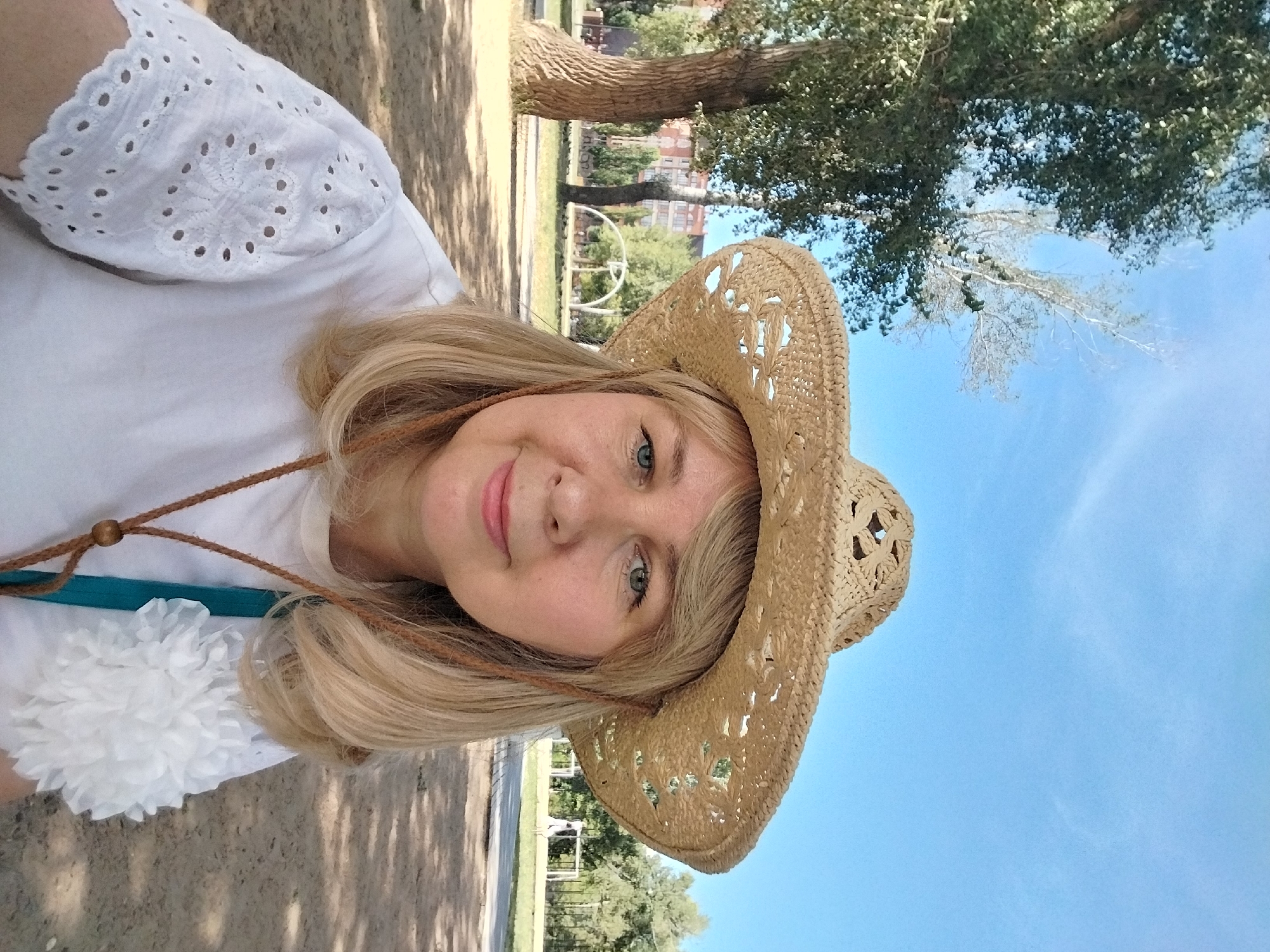 A blonde woman in a straw cowboy hat and a white shirt with eyelet sleeves poses for a selfie outdoors. She is wearing a turquoise bag strap and a brown leather cord necklace with a wooden bead. The background shows trees, a building, and a blue sky with some clouds. The location is the city of Orenburg, Russia.