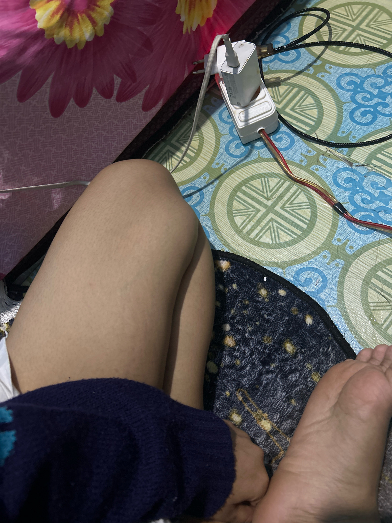The image shows a close-up view of a person's legs and feet, along with some electrical objects and a patterned surface. The person is wearing a dark blue knitted garment around their lower torso. Their legs are bare, and the skin appears to have some minor texture. The feet are resting on a dark blanket with a speckled pattern.
In the background, there is a white electrical power strip plugged into an outlet, with a white phone charger and its cable attached. Several other wires, including one with red and yellow insulation, are also visible, connected to the power strip. The power strip and wires are placed on a light blue and green patterned surface, possibly a mat or a floor covering, with a circular design. To the left, a portion of a pink and yellow floral pattern is visible, possibly from a curtain or bedding.
The scene suggests an indoor setting, possibly a bedroom or a living area, given the presence of soft furnishings and electrical devices. There are no clear indications of the time of day or weather conditions. The composition focuses on the personal space of the individual and their immediate surroundings. No visible text is present in the image.