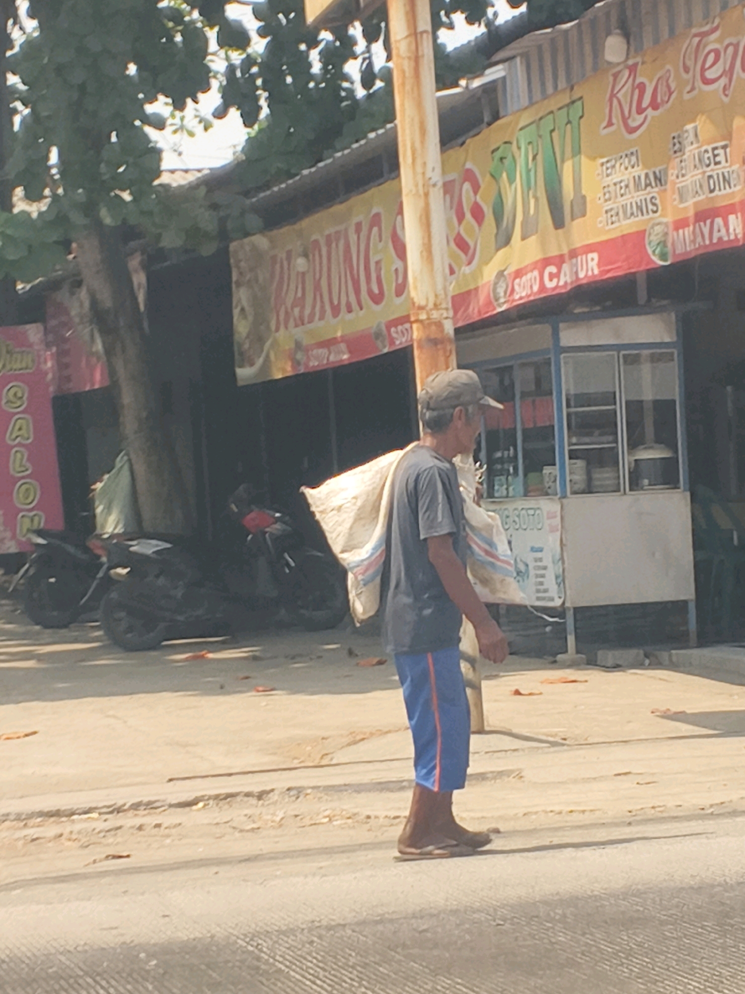 A medium shot captures a man walking on a dusty road in Sutamaja, Indonesia, carrying a large sack over his right shoulder. He is wearing a gray t-shirt, blue shorts with an orange stripe, and brown sandals. The man is walking away from the camera, towards a row of small shops. One shop has a yellow banner with red text that appears to advertise food items. Two motorcycles are parked in front of the shops. The ground is unpaved and sandy, with scattered debris. The background shows foliage and the roofs of buildings. The lighting suggests a bright, sunny day.