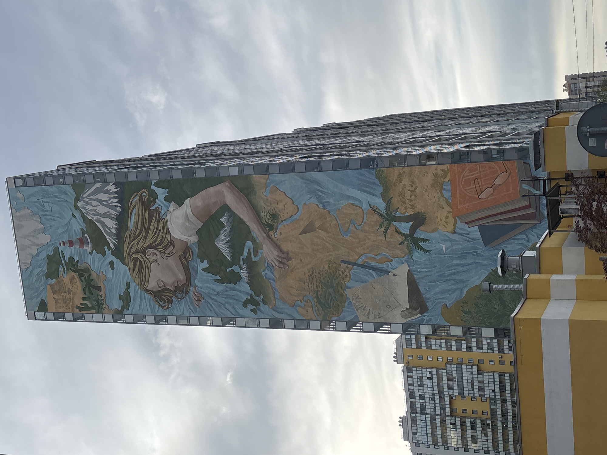 The image displays a towering building adorned with a large, vibrant mural covering a substantial portion of its side. The mural depicts a woman with long, flowing blonde hair, her head tilted, and her eyes seemingly closed in a serene or contemplative state. She appears to be emerging from or immersed in water, which surrounds a world map.
The map features various landmasses in earthy tones against blue waters. To the left of the woman's head, a lighthouse stands in the water near a mountainous or icy region. Her right arm extends across the map, with her hand resting on a landmass. Further to the right on the mural, a palm tree is visible on a different landmass, suggesting warmer climates. Additional elements in the mural include a sailing ship or boat, several stacked books, and a pair of reading glasses, along with what resembles a rolled map or compass.
The building itself, visible above and below the mural, has a stepped or layered architectural style with numerous windows or balconies. Below the mural, the building transitions to a section with distinctive yellow and white vertical stripes. In the lower right background, another tall building with a similar modern design and yellow accents can be seen.
The sky is overcast with soft, diffused light, indicating a cloudy daytime setting. No discernible text is visible on the mural or the buildings. The overall impression is one of artistry integrated into urban architecture, with themes of travel, knowledge, and exploration conveyed through the mural.