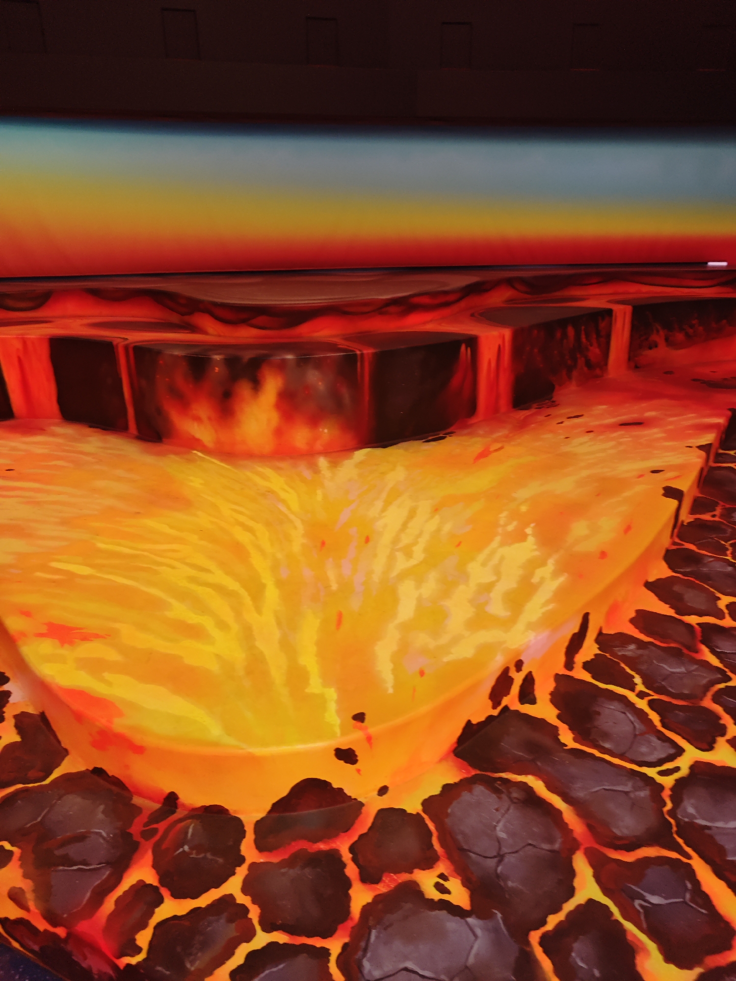 The image depicts a visually striking, stylized representation of flowing lava. The lava is rendered in vibrant shades of orange, yellow, and red, with darker, cracked volcanic rock formations in the foreground. The lava appears to be cascading over dark, rectangular structures. Above the lava, there is a band of gradient colors transitioning from deep red to yellow and then to a pale blue, suggesting an atmospheric effect or a stylized sky. At the very top of the image, a dark horizontal band with subtle rectangular shapes could represent buildings or structures in the background.
This image does not depict any specific location in the city of Eindhoven, Netherlands, nor does it contain any people, actions, or interactions. It is an artistic or illustrative representation of lava.