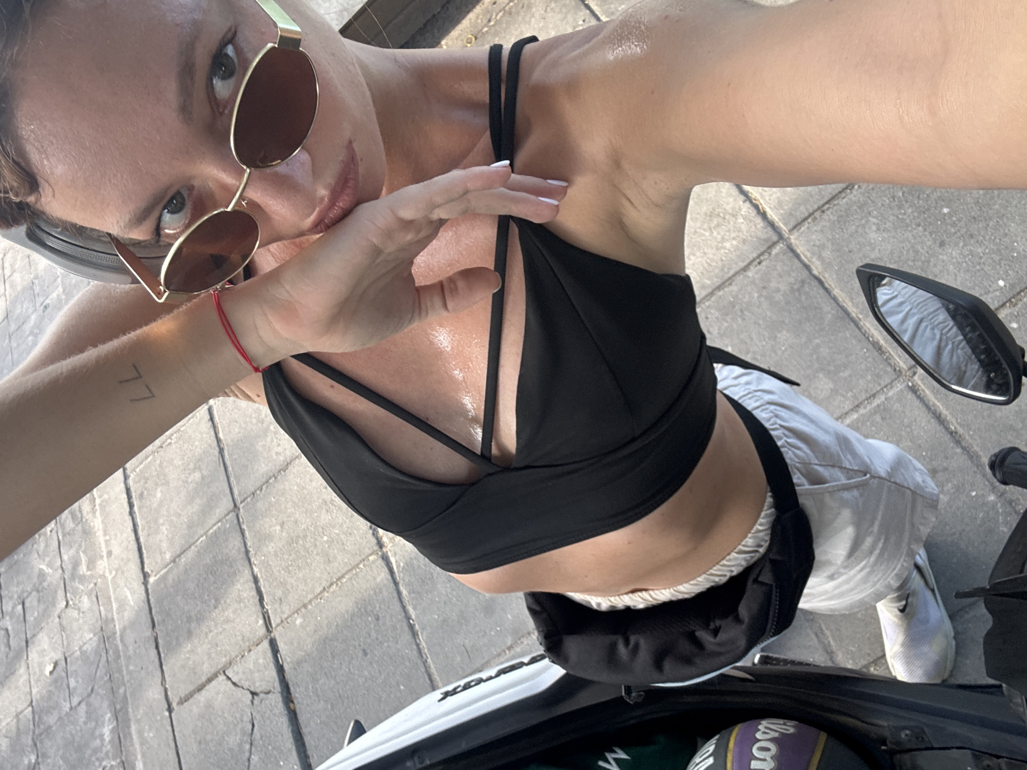 In a high-angle, selfie-style shot, a woman with light brown hair, wearing sunglasses and a black crop top, rests her hand on her chin. She is positioned on a scooter, as indicated by the handlebar and mirror visible in the lower right corner of the frame. The background consists of grey pavement with visible grout lines. A basketball with the brand "Wilson" is partially visible in a compartment on the left side of the scooter. The woman has a red string bracelet on her left wrist and a tattoo on her left forearm. The lighting suggests it is daytime, with sunlight casting shadows on the ground. The environment appears to be an outdoor urban setting, possibly a street or parking area in the Ko Samui District of Thailand.