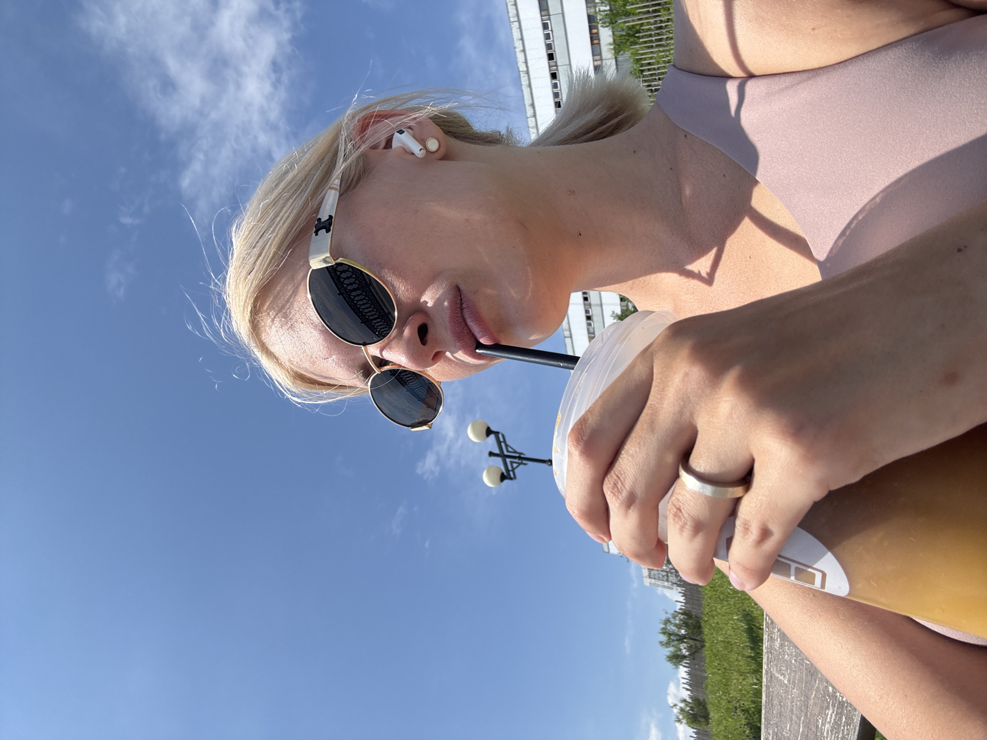 A blonde woman with sunglasses is drinking from a clear plastic cup with a straw. She is wearing a pink top and a ring on her left hand. She has a white wireless earbud in her right ear. The background shows a blue sky with clouds, a lamppost, trees, a fence, and a building. The scene appears to be outdoors in a park or public area.