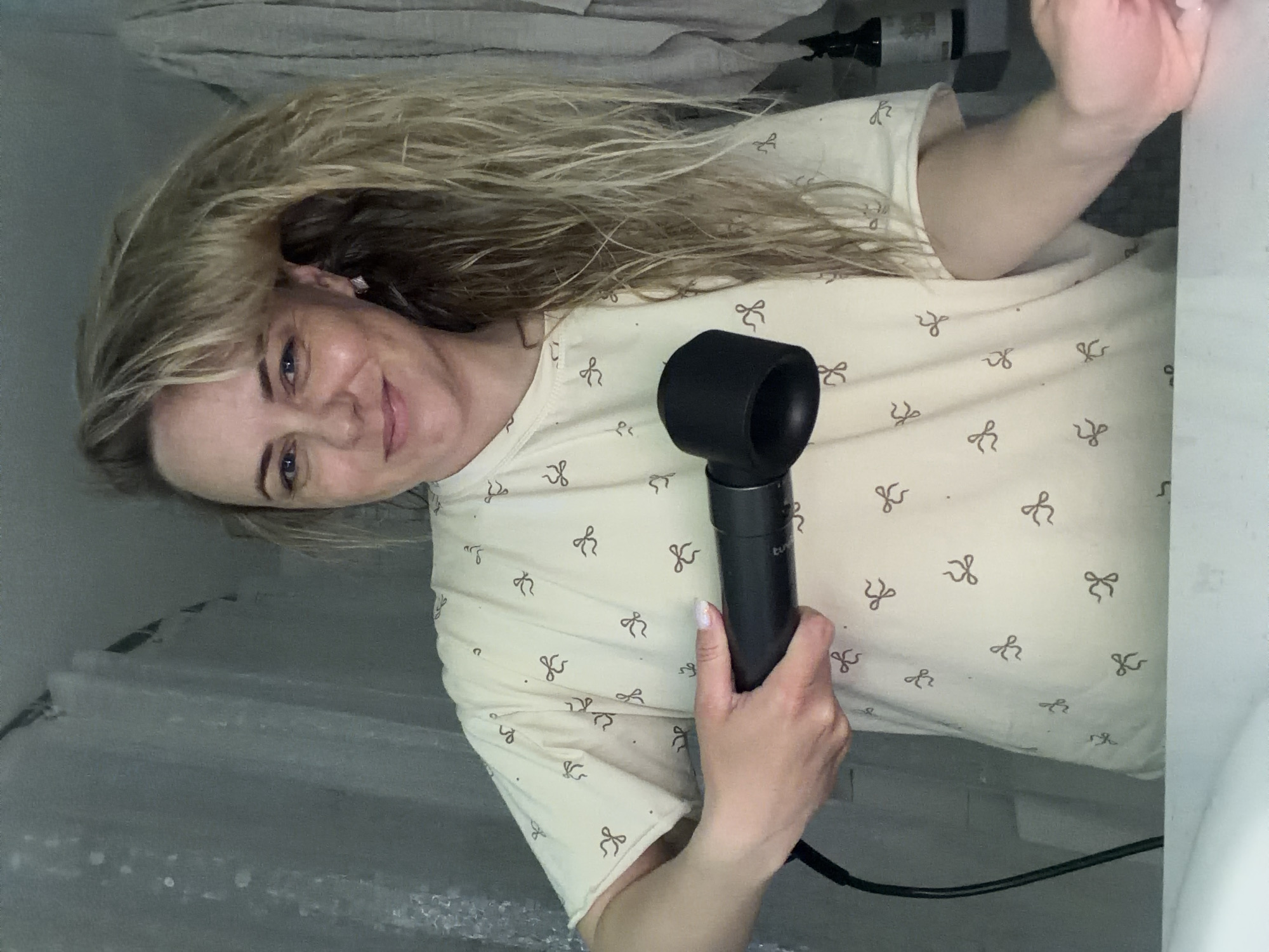 The image, oriented sideways, depicts a woman with light, wavy hair and blue eyes, smiling gently at the camera. She wears a cream-colored t-shirt patterned with repeating dark brown bow ties. In her right hand, she holds a distinct black hairdryer, its cord extending downwards. Her hair appears somewhat damp, suggesting she is either preparing to dry it or has recently washed it.
The setting is an indoor space, likely a bathroom, indicated by a light-colored fabric or towel behind her head and bottles visible on a surface to her left. The lighting is artificial and appears somewhat dim. The woman's expression is calm and pleasant. There is no clearly legible text within the image; the OCR output consists of fragmented characters and symbols, which are likely misinterpretations of the shirt's pattern and background textures. While the provided location context is Votkinsk, Russia, no specific visual cues within the image itself confirm this detail.