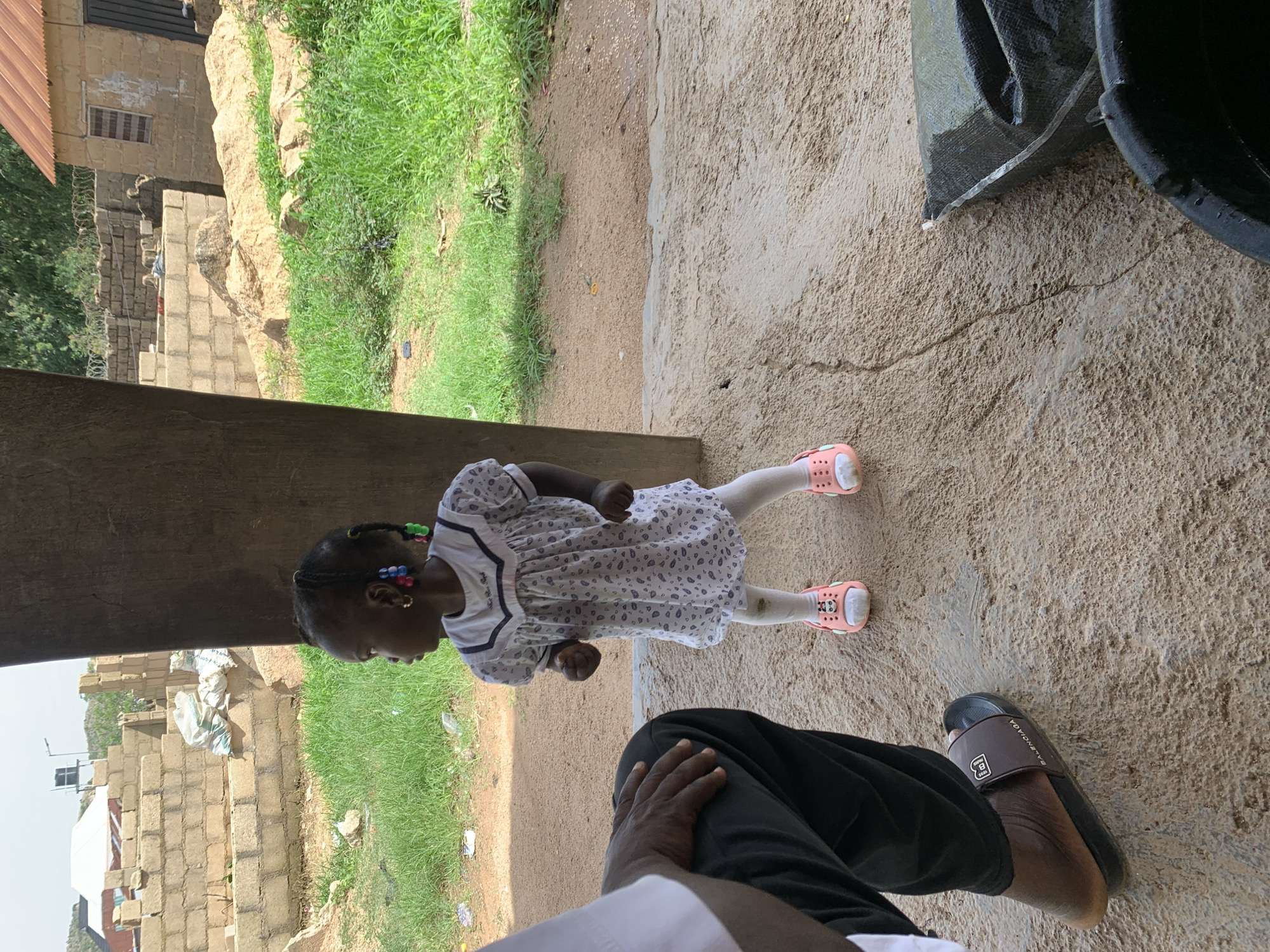 In Jos, Nigeria, a young African girl stands on a sandy patch of ground near a large wooden post, wearing a white dress adorned with a subtle pattern, white leggings, and pink sandals with cartoon characters. Her hair is styled in braids, decorated with colorful beads. To her left, a person's legs and torso are visible, clad in black trousers and a sandal with the "Balenciaga" logo. The background reveals a construction site with brick walls, piles of sand, and scattered green grass. A portion of a building with a reddish-brown roof is also visible. The scene suggests a rural or developing urban setting.