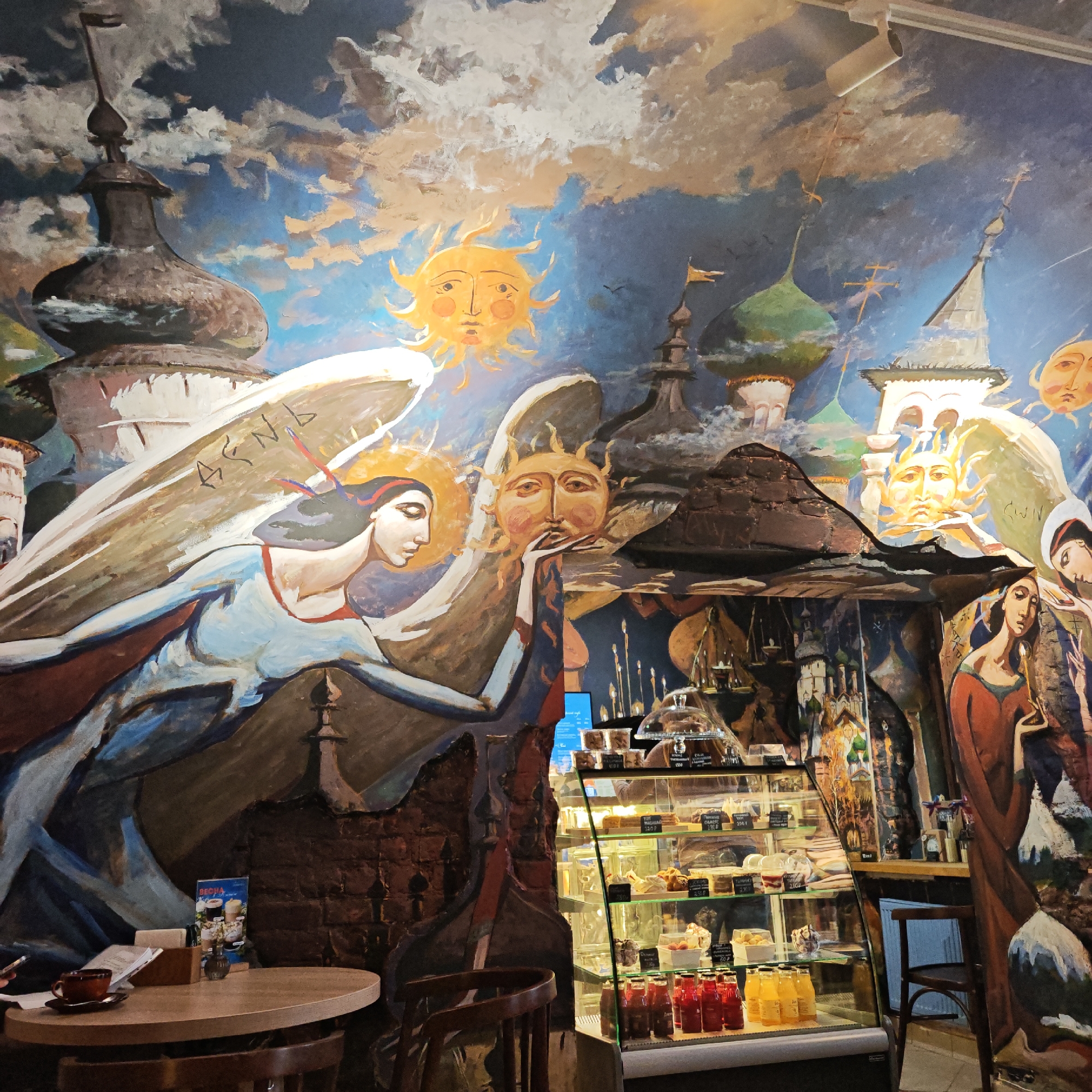 The image shows the interior of a cafe in Rostov, Russia. The walls are adorned with a large, mural-style painting featuring angels, stylized suns, and towers reminiscent of Russian architecture. A wooden table with a cup and a small card stand in the foreground. A refrigerated display case containing cakes and drinks stands against a section of the painted wall. A wooden chair sits beside the display case, creating a somewhat cramped space in the cafe.