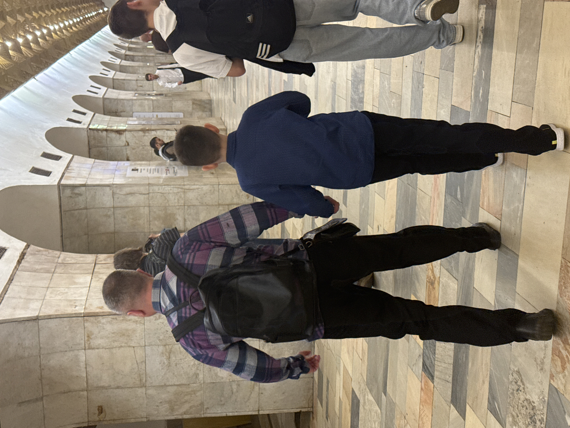 The image shows a group of people walking in a subway station in Moscow, Russia. The station is characterized by its arched ceilings and tiled walls. The floor is paved with rectangular tiles in various shades of beige and gray.
In the foreground, a man with a shaved head is walking, wearing a purple and gray plaid shirt, black pants, and black shoes. He also has a black backpack on. Next to him is a child in a blue shirt and black pants, also walking in the same direction. Above them is a person in a white shirt, black vest, and gray sweatpants.
In the background, other people are walking further away. The lighting is diffused, creating a somewhat muted atmosphere. The subway station’s architecture is visible, showing a series of arched alcoves along the walls.