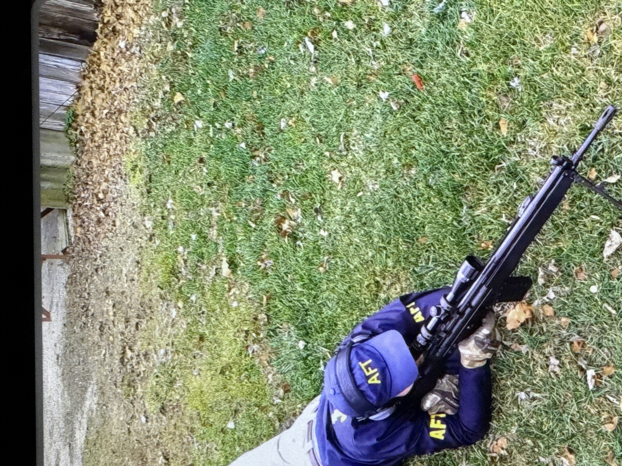 An individual, wearing a blue cap with "AFT" visible and a dark blue jacket with yellow "AFT" lettering, is lying prone on a grassy area. The individual is holding a rifle equipped with a scope. The environment appears to be an outdoor setting with fallen leaves scattered across the grass, suggesting an autumn season. A wooden fence is visible in the background, along with some structures. The scene is set in the city of Cahul, Moldova.