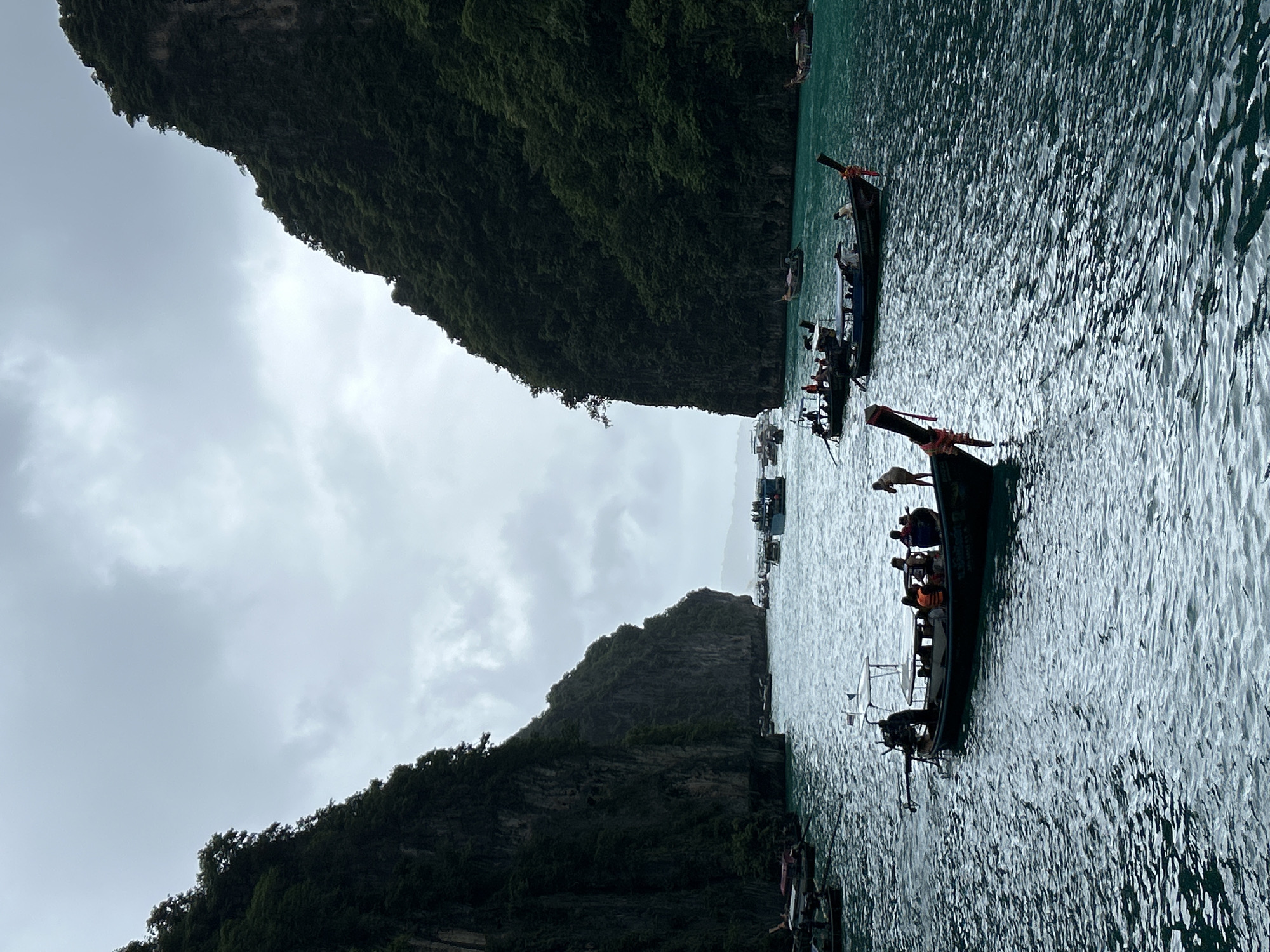 This vertical, eye-level shot captures a scenic view of a bay in Ao Nang, Thailand, characterized by towering, lush green karst cliffs on either side. The water is a deep turquoise, with small waves rippling across its surface, reflecting the overcast sky. Several long-tail boats, traditional Thai vessels with distinctive wooden prows, are scattered throughout the bay. One boat in the foreground is filled with passengers, some wearing life vests, and a person stands at the stern near the engine. Another similar boat is slightly further back, also carrying people. In the middle ground and background, more boats of various sizes are visible, some appearing to be tour boats with multiple passengers. The scene conveys a sense of a popular tourist destination, with the boats suggesting activity and exploration within this striking natural landscape. The weather appears to be cloudy, creating a soft, diffused light that contributes to the slightly moody atmosphere of the image.