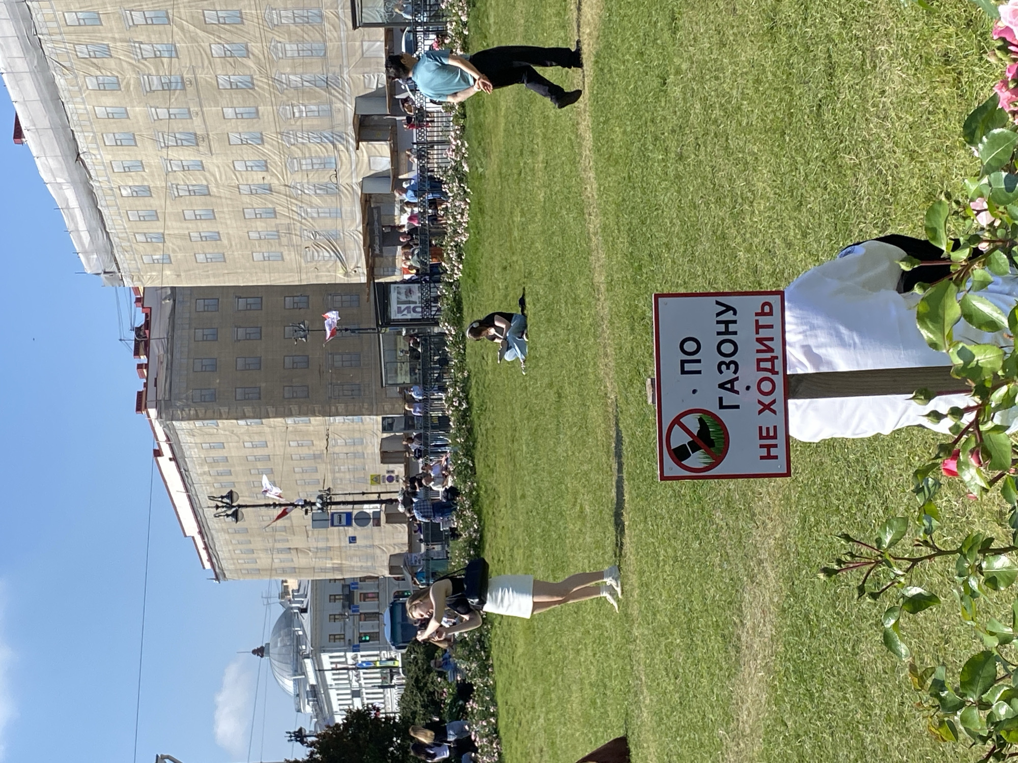 A woman in a white skirt and black top stands in a park, holding a phone to take a picture. She is on a green lawn, and in the background, there are buildings, some covered in scaffolding. A red and white sign with Russian text and a prohibitory symbol is visible in the foreground. Several other people are scattered across the lawn, some sitting and others walking. The sky is blue with some clouds. The scene is likely in Saint Petersburg, Russia, given the architectural style of the buildings and the Russian signage.