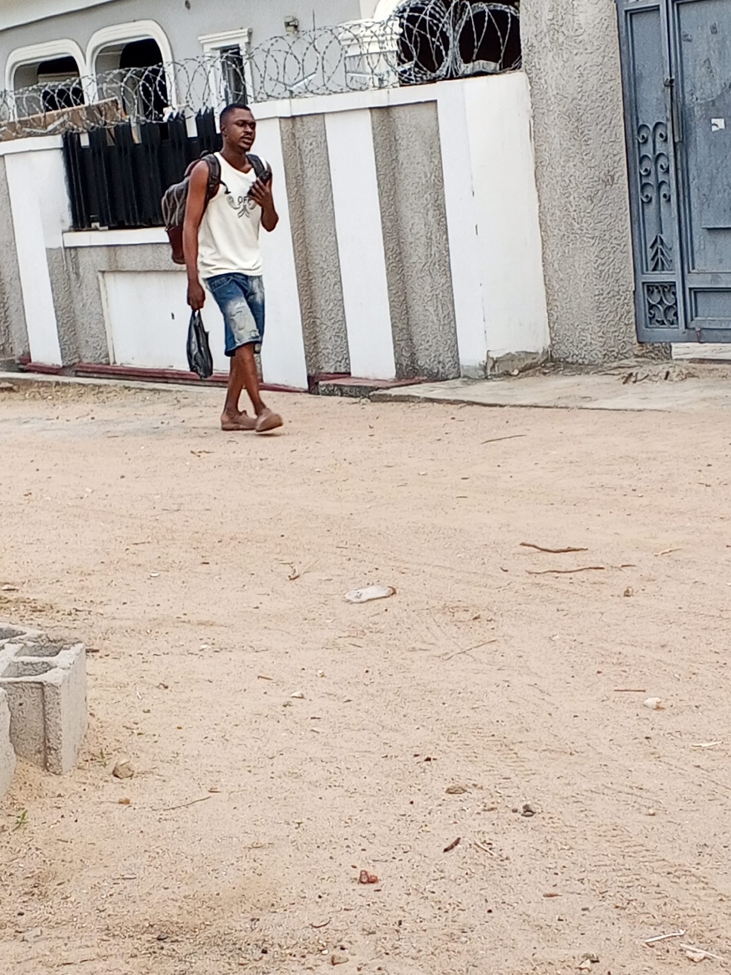 The photo shows a young man walking on a dirt road in Adonkolo, Nigeria. He is wearing a white tank top, denim shorts, and brown sandals. He has a backpack on and is holding a dark bag in his right hand. The street is unpaved, with a sparse scattering of debris. There is a building with a white and gray facade behind him, topped with barbed wire. A gray door is on the right of the building. A low concrete block is visible in the lower left corner of the image.