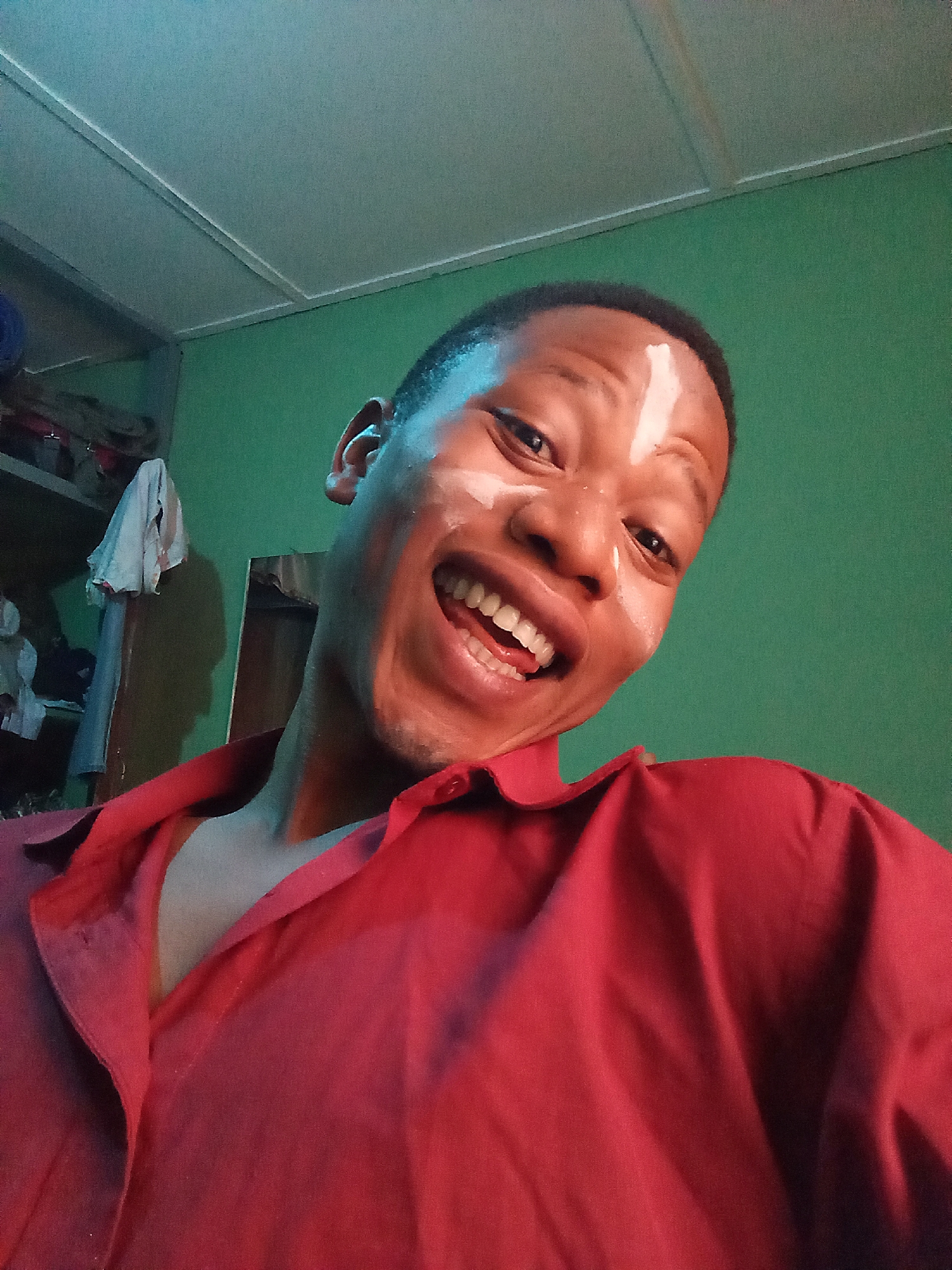 A young African man is smiling broadly, revealing his teeth. He has white markings on his forehead and cheek. He is wearing a red shirt with the collar open. The background is green, and there is a metal shelf with objects on it to the left. The location is indoors. There is no information given about the city of Oshogbo, Nigeria.