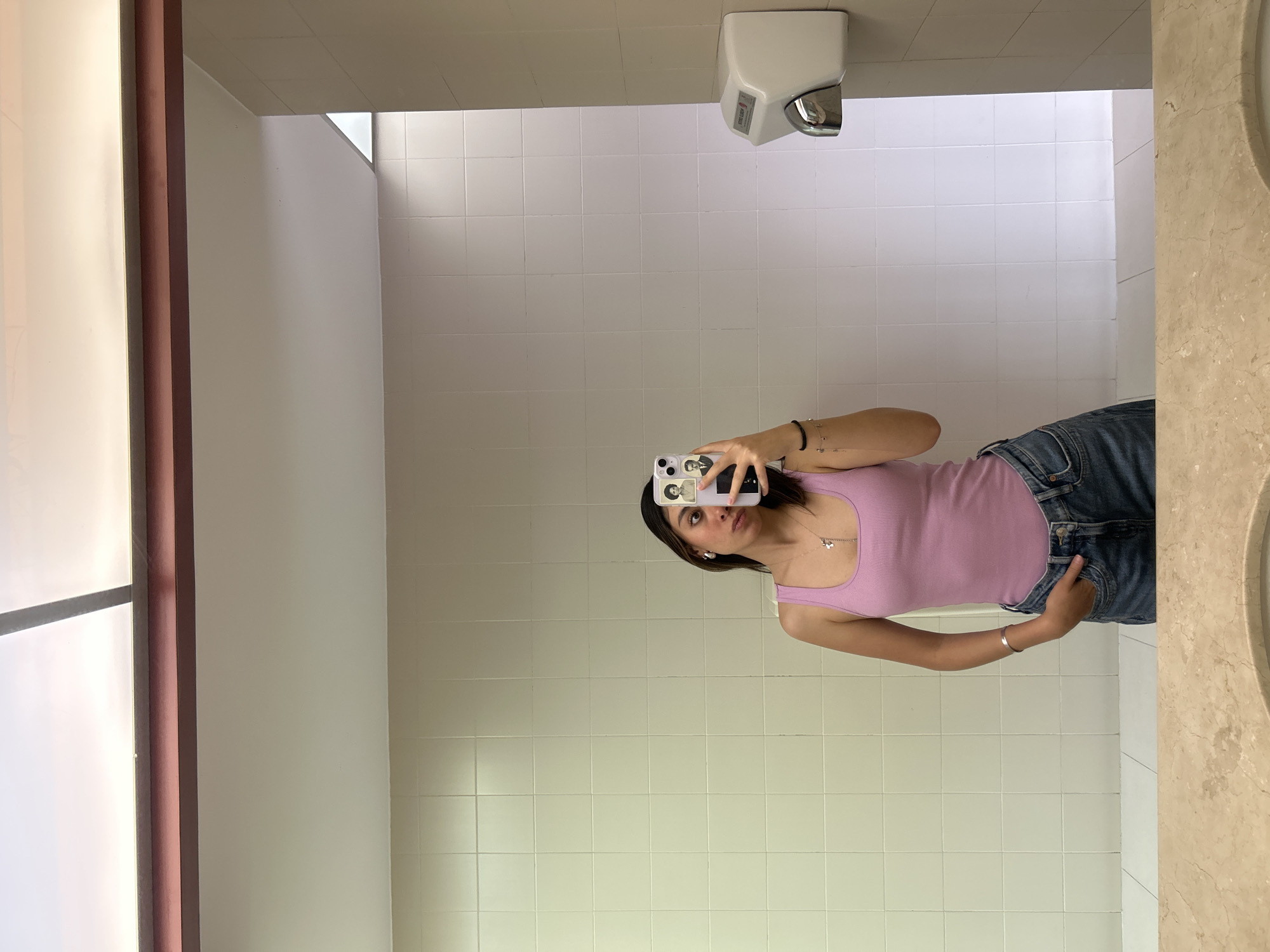A young woman is captured in a mirror selfie, standing indoors against a backdrop of white tiled walls. She is wearing a light purple ribbed tank top and blue denim jeans, with her left hand casually tucked into her front jeans pocket. Her right hand holds a white smartphone with a decorative case, partially obscuring her face as she looks directly into the camera lens. She has dark hair, a silver bracelet on her left wrist, and a delicate necklace with a small charm.
The setting appears to be a public restroom or similar facility. A white hand dryer is mounted on the wall in the upper right portion of the image. The ceiling is a plain, light color. To the far left, a large, vertical rectangular light source, possibly a window with frosted glass or a large light panel, provides illumination, creating bright highlights. A brown or maroon vertical beam or pipe runs alongside this light source. The floor is not visible, but the wall tiles extend downwards. The light seems to be natural or bright artificial. No discernible text is clearly legible on the hand dryer or the phone case.