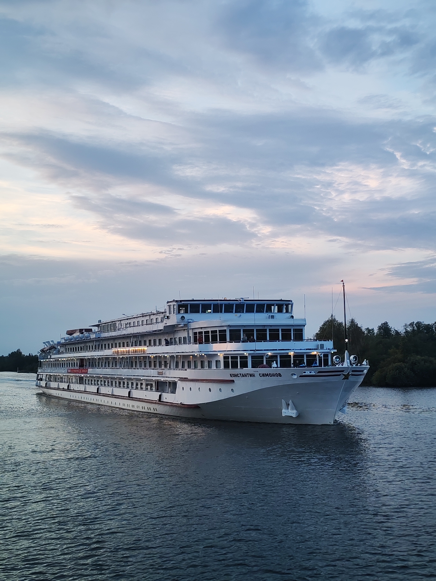 A white passenger ship, named "КОНСТАНТИН СИМОНОВ" in Cyrillic, is navigating a body of water, likely a river, under a cloudy sky. The ship is large, multi-decked, and appears to be in motion, indicated by the ripples on the water's surface. The scene suggests a leisurely cruise, possibly during dawn or dusk, given the soft lighting and muted colors of the sky. The ship is white with red accents along its hull and decks. The background consists of a dark, tree-lined shore on the right, partially obscured by the ship. The overall impression is one of peaceful travel in a natural setting. This scene is consistent with the views found along the banks of the Vytegra River in Vytegra, Russia, a town known for its historical significance and its connection to waterways like the Onega Canal.